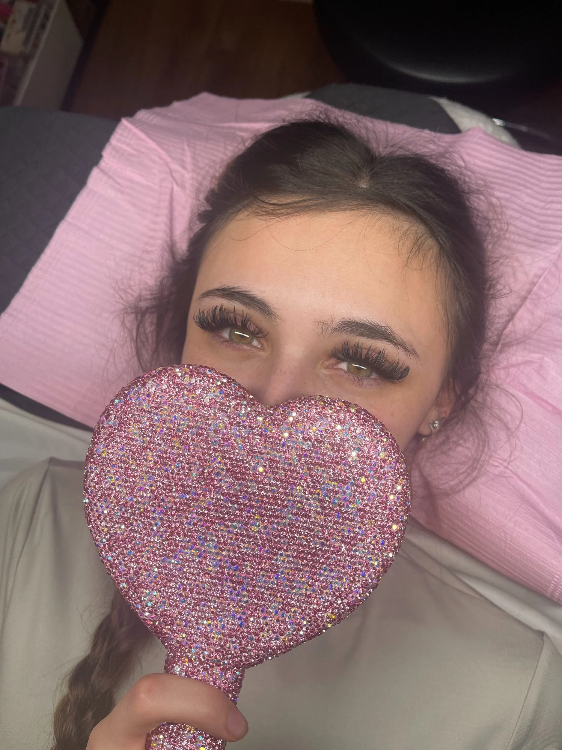 Young woman with long eyelashes lying on a pink pillow, holding a large pink glittery heart-shaped mirror in front of her face, partially covering her mouth and nose.