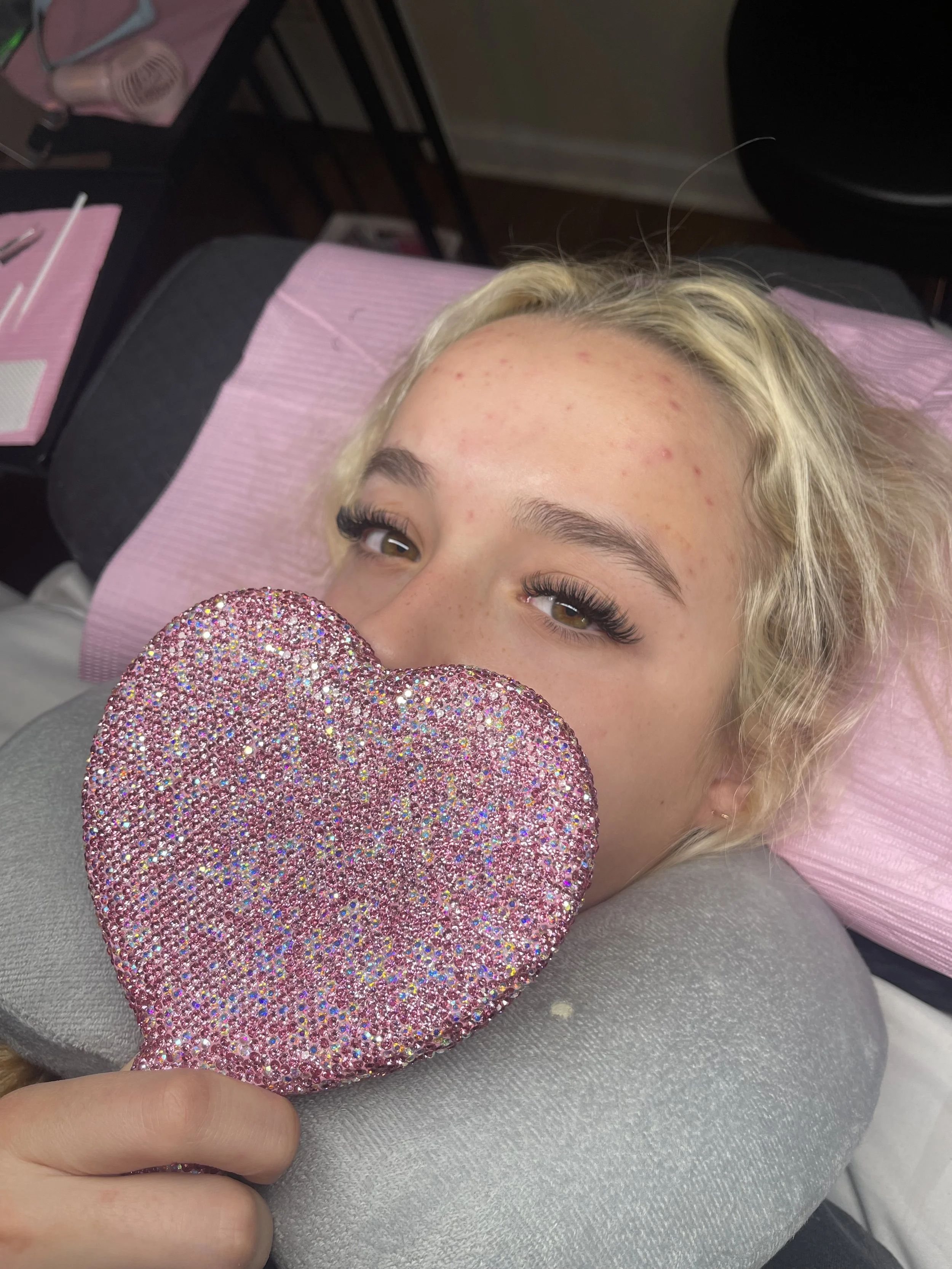 Young woman with blonde hair, lying on a treatment bed with a pink pillow, holding a sparkly pink heart-shaped cushion, showing her face with eye makeup and some acne on her forehead.