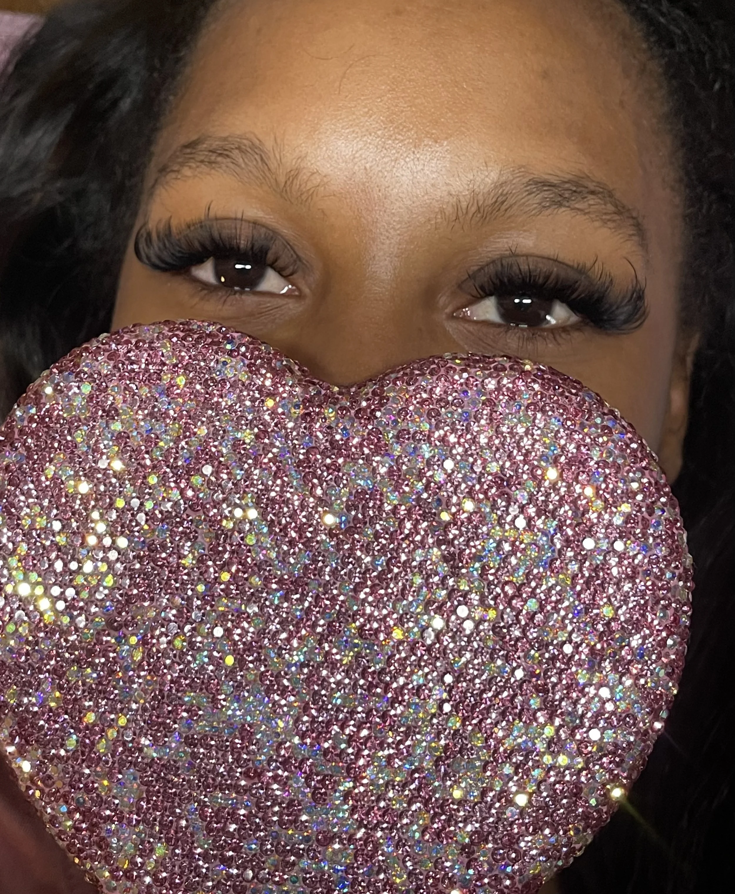 A woman with long eyelashes holds a large, sparkling, pink, rhinestone-encrusted heart close to her face, covering her nose and mouth, with only her eyes visible.