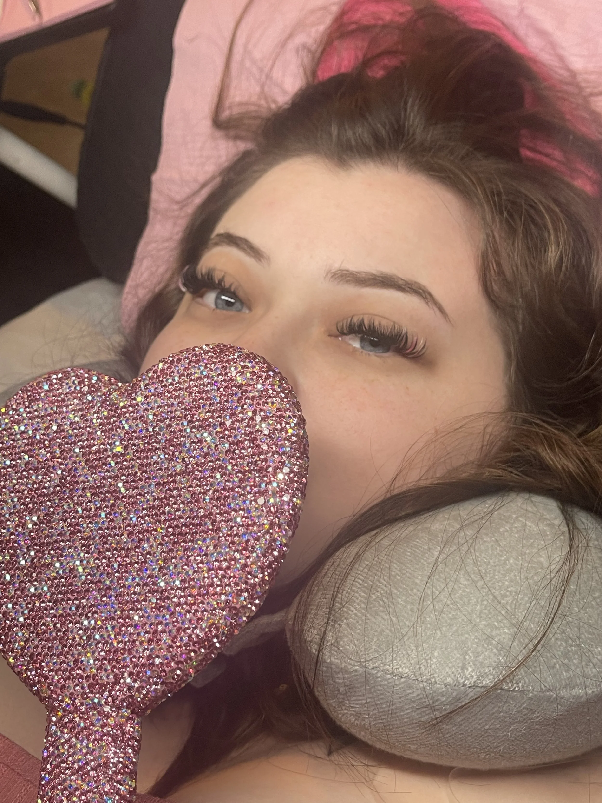 A young woman with long brown hair and blue eyes lying on a pillow, holding a sparkly pink heart-shaped object partially in front of her face.