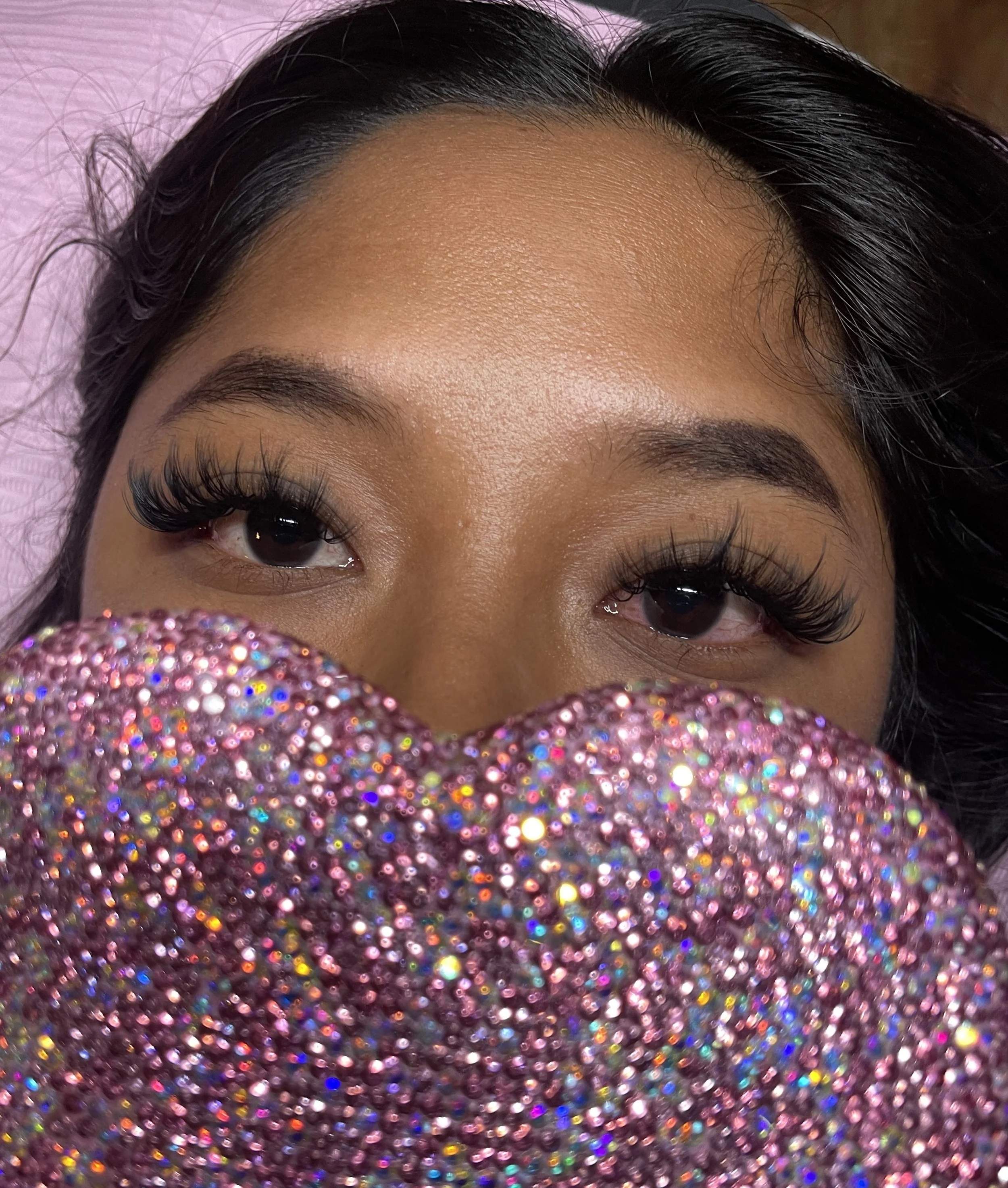 Close-up of a woman's face with her eyes visible, partially covered by a glittery, pink, rhinestone-encrusted object.
