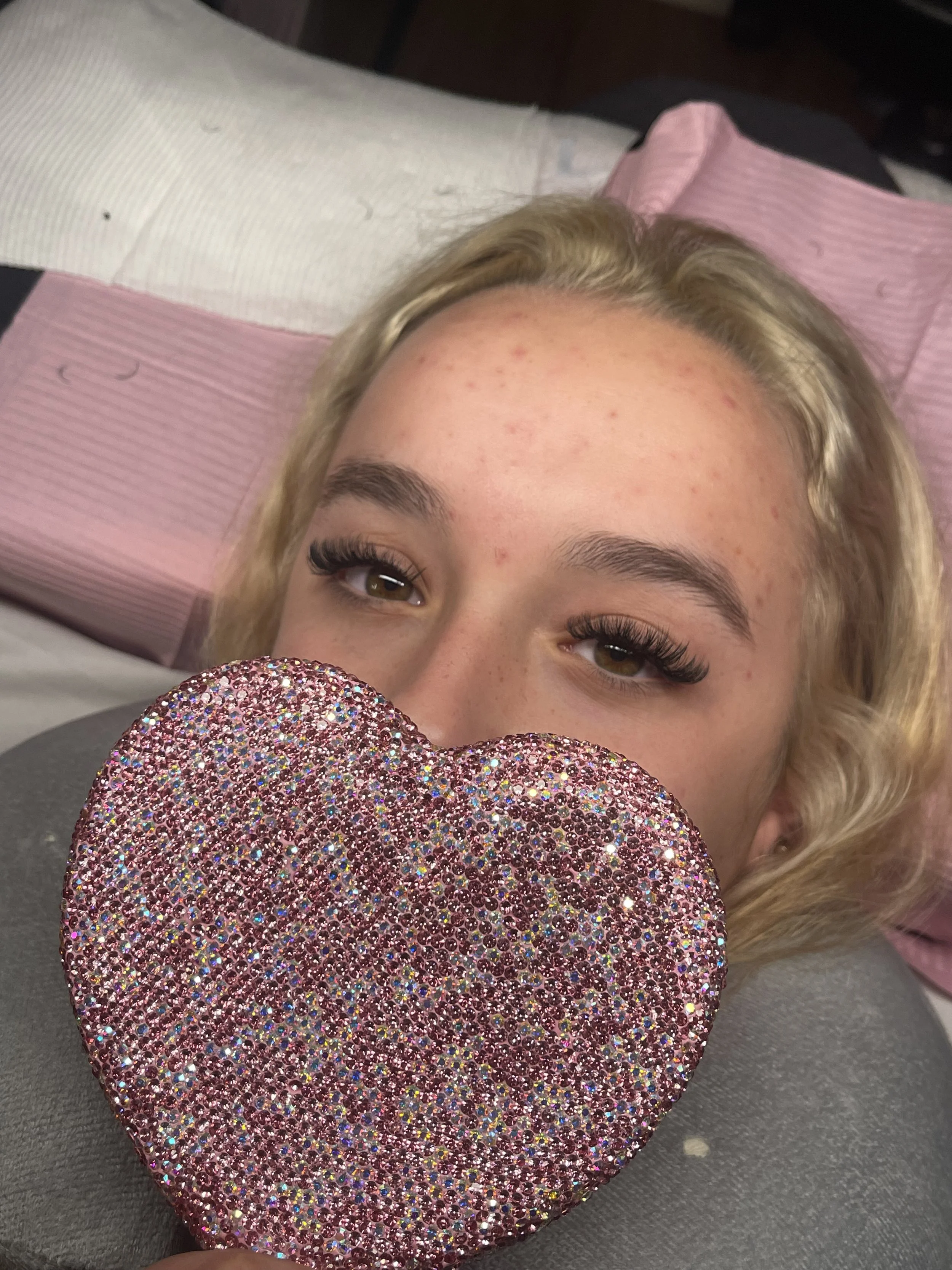 A young woman with blonde hair and thick false eyelashes lying down, holding a large pink, sparkling heart-shaped object close to her face, with a pillow and blankets in the background.