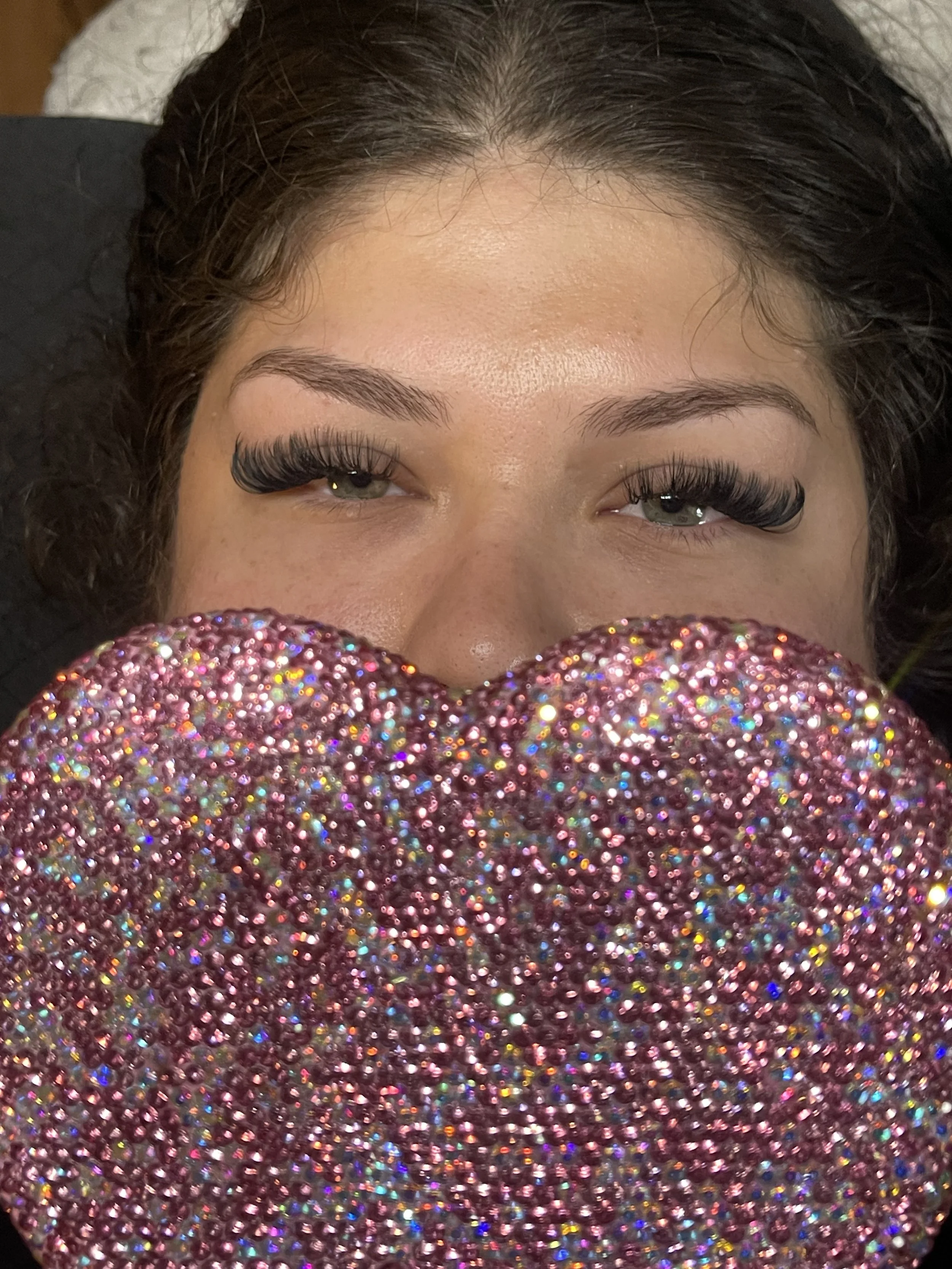 Close-up of a woman's face with eye makeup, holding a large, glittery pink heart in front of her mouth, with only her eyes and part of her nose visible.