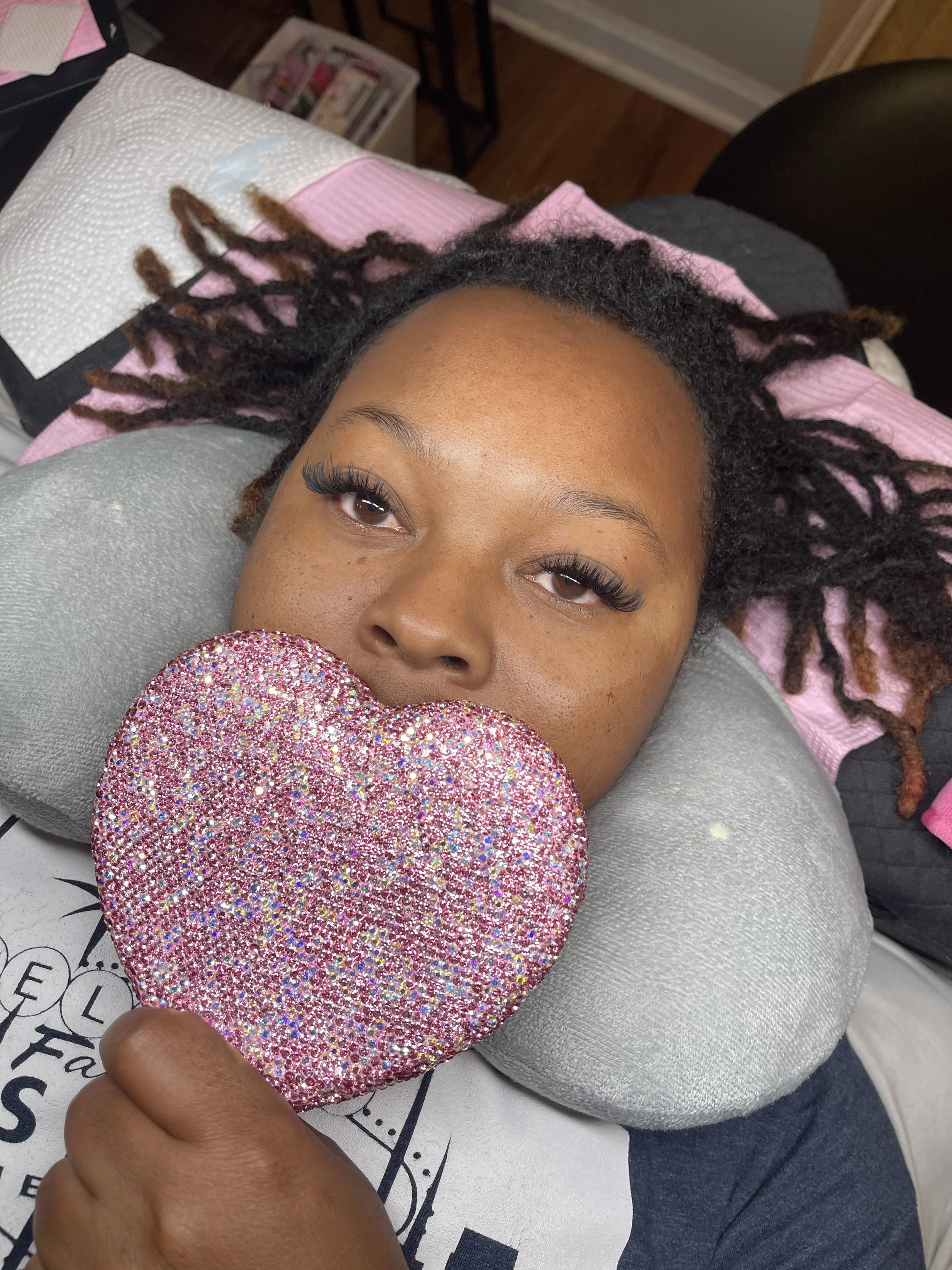 A woman lying on a pillow, holding a pink and multicolored rhinestone heart, with her eyes partially closed.
