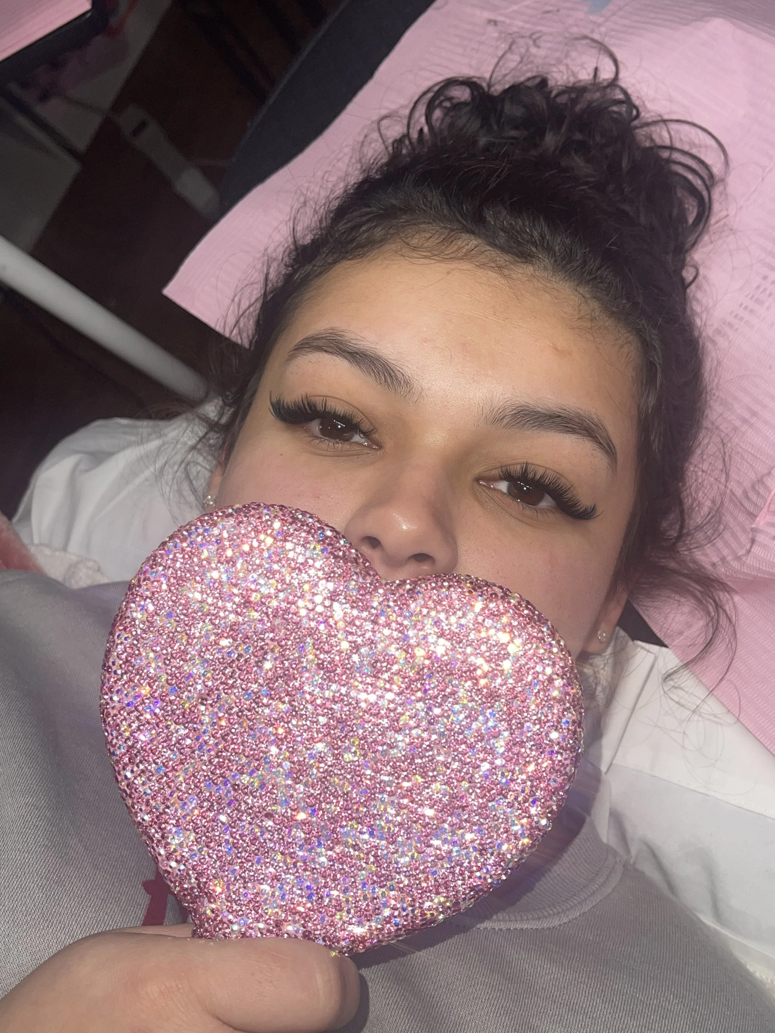 A woman with curly hair and eyelash extensions lying down, holding a large sparkly pink heart-shaped object in front of her face.