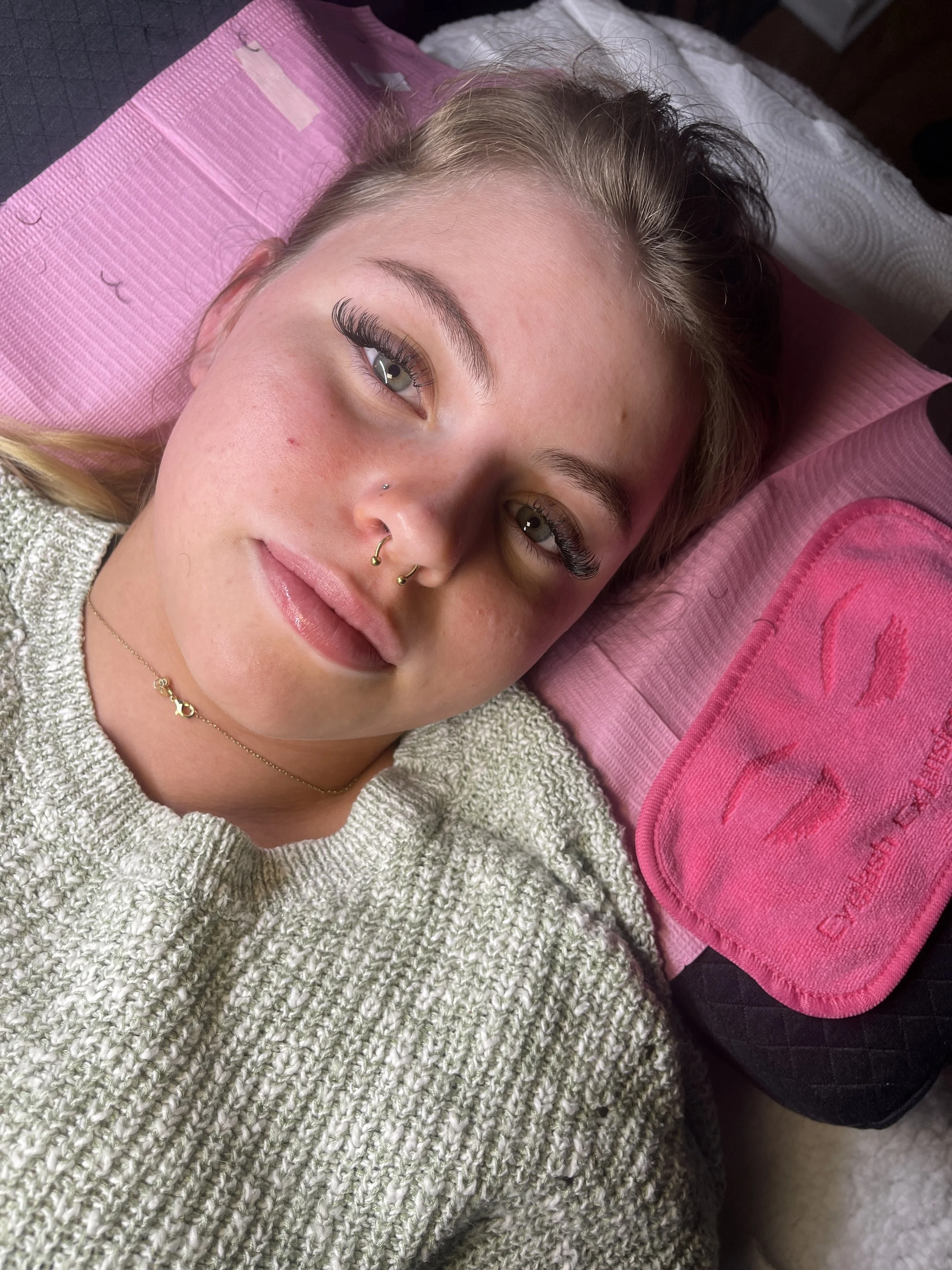 A young woman with blonde hair, lying down on a pink and black dental chair with a pink headrest and a pink dental mouthguard pillow, wearing a knitted sweater and jewelry, looking at the camera.