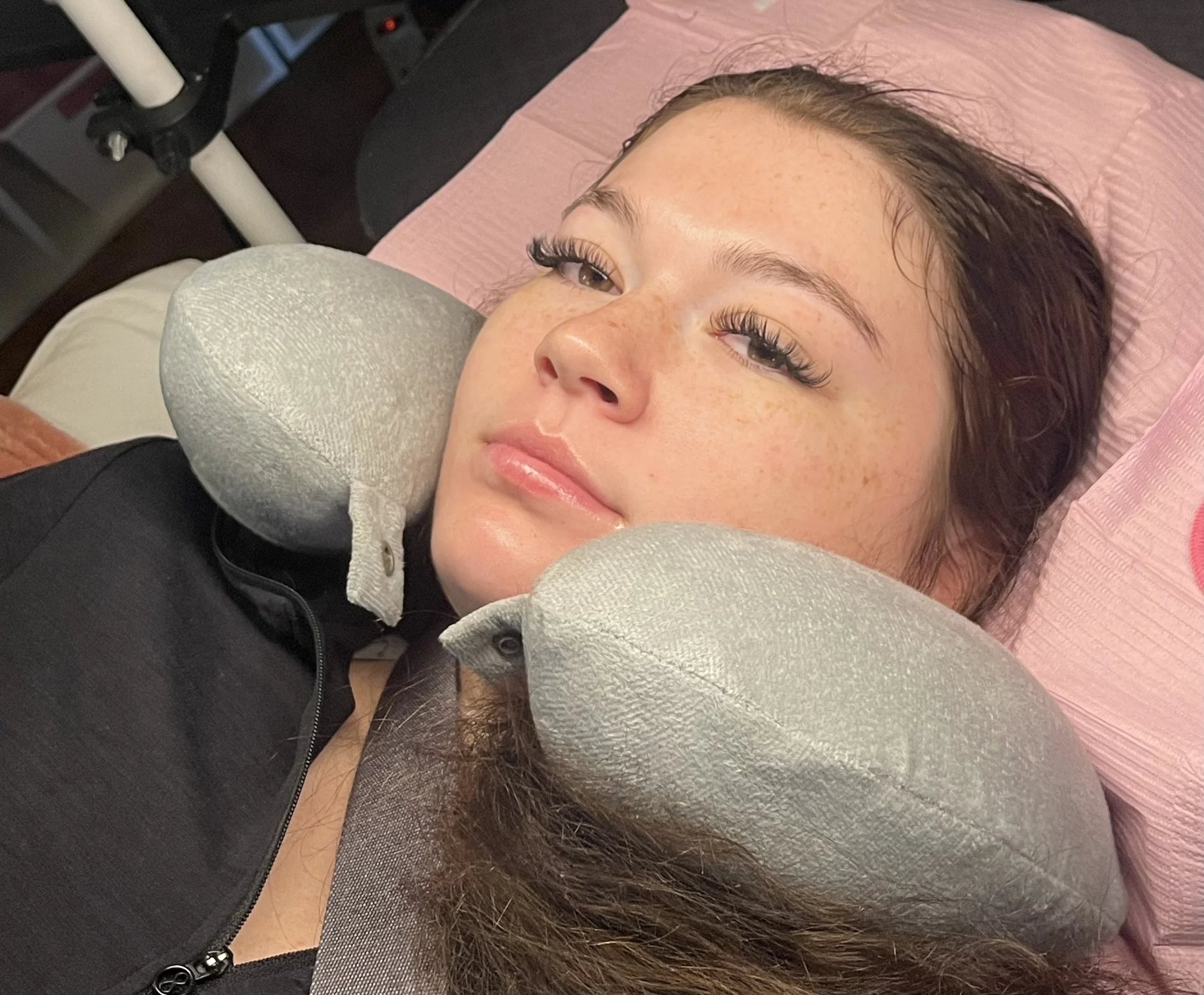 Young woman with curly brown hair lying on a pink pillow, wearing a black shirt, with two gray neck pillows around her neck and shoulders, in a medical or spa setting.