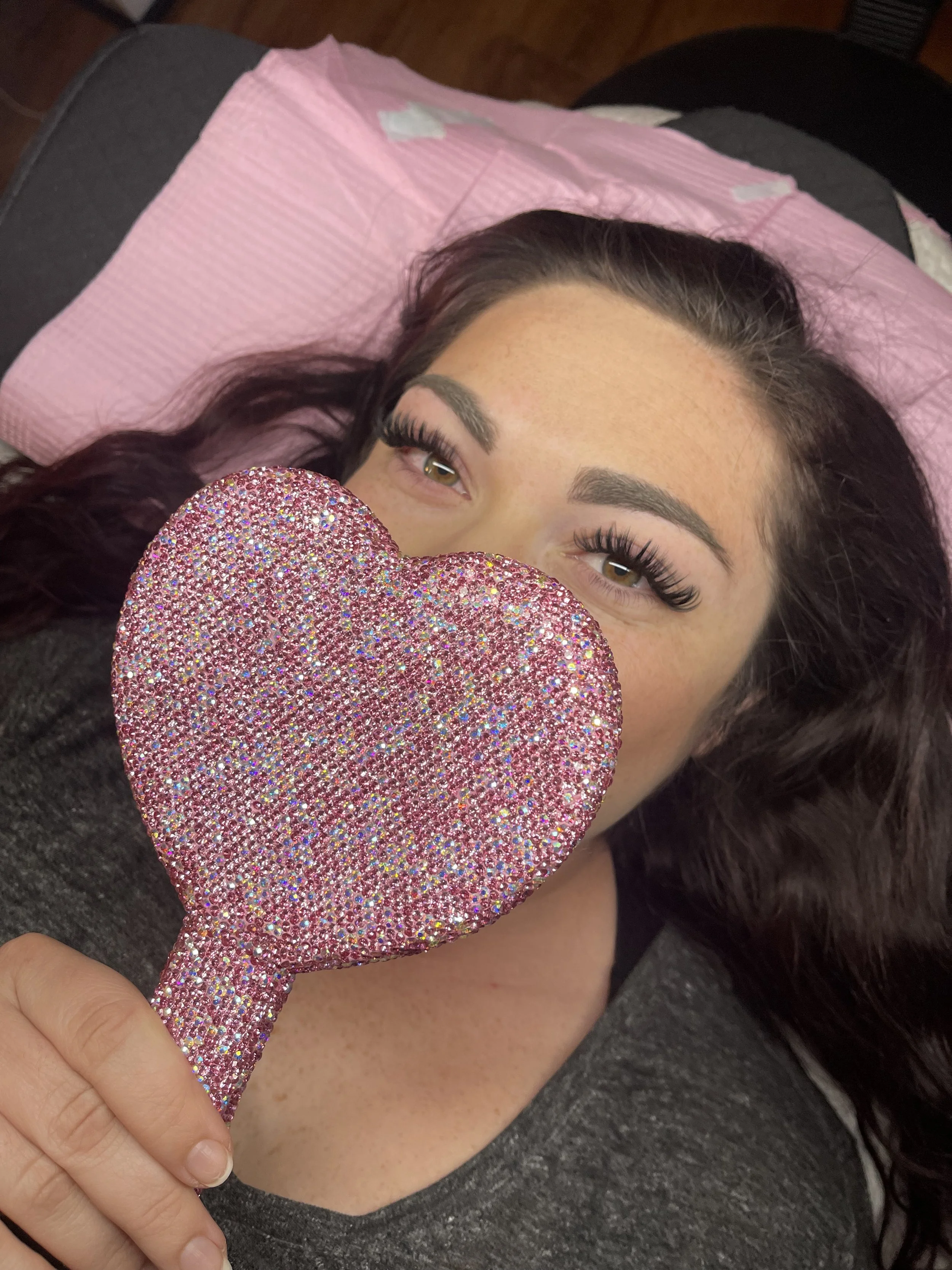 A woman with dark hair and makeup lying on a pink pillow, holding a sparkly pink heart-shaped mirror in front of her face.