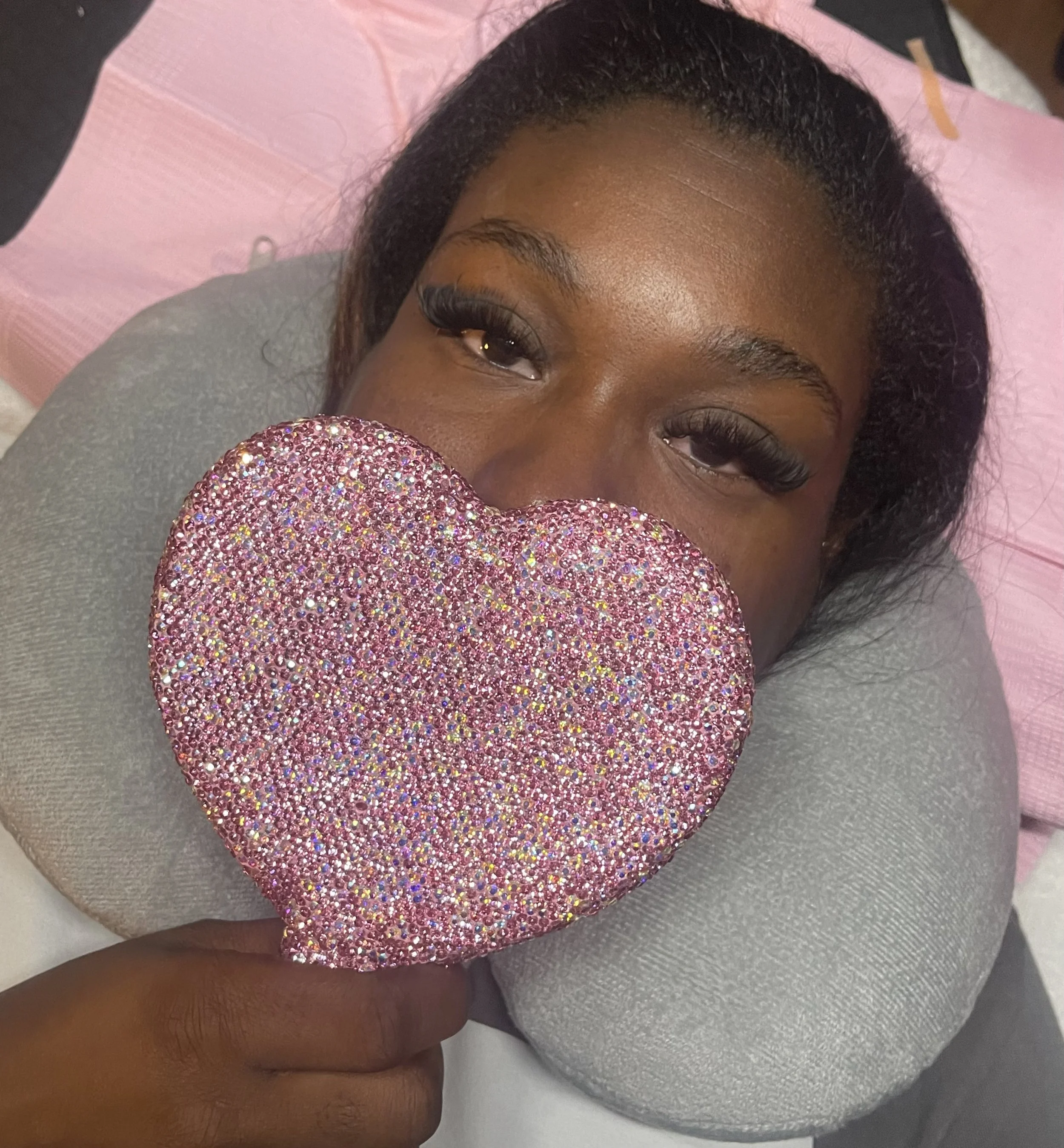 A woman lying down, holding a sparkly pink heart-shaped object in front of her face, with her eyes visible.
