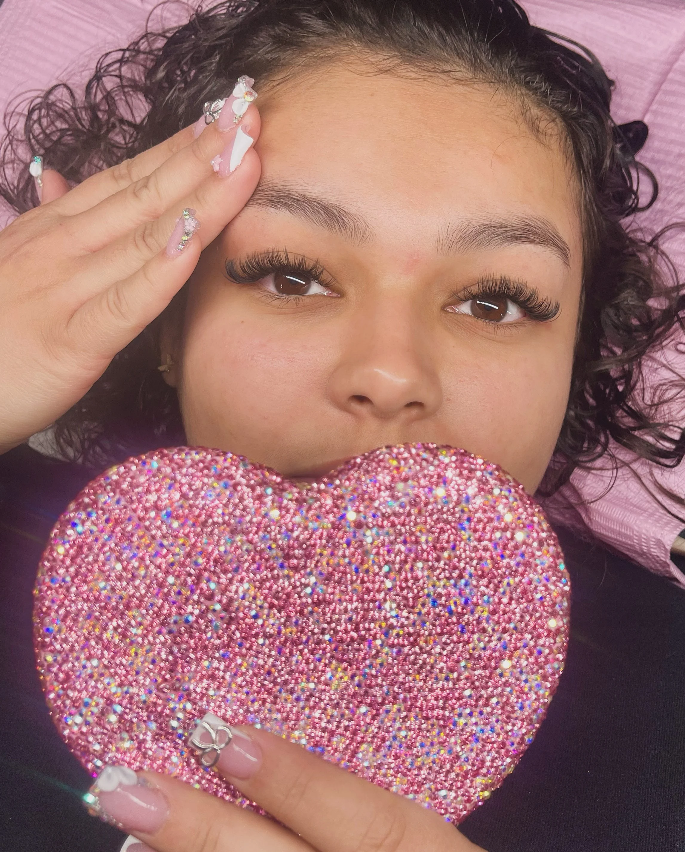 A woman with curly hair lying down, holding a pink, sparkly, heart-shaped object in front of her mouth, with her left hand touching her forehead.