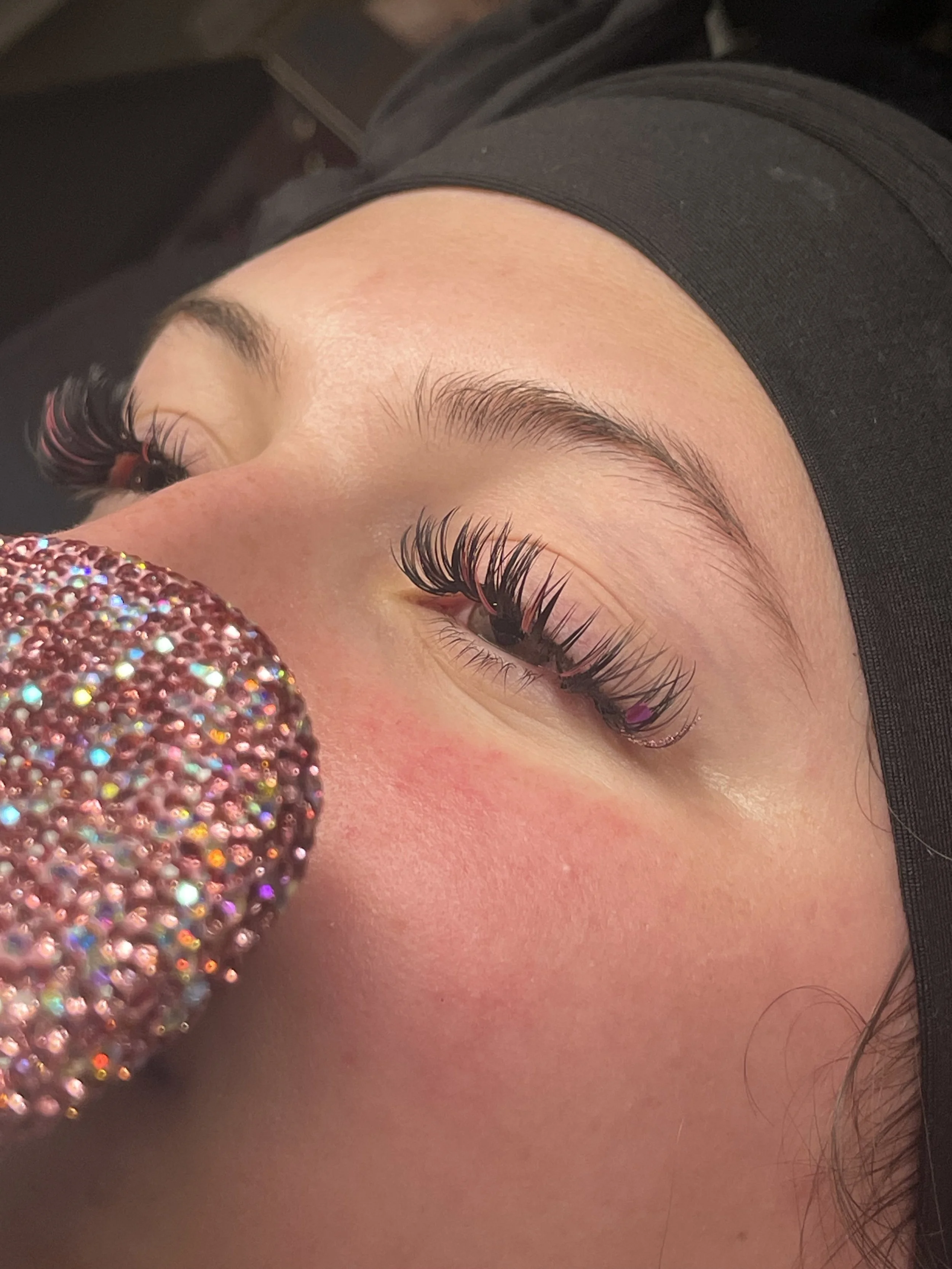 Close-up of a woman's face showing her eye with long, curled, and voluminous eyelashes, with a black headband, and pink blush on her cheek. A sparkly, rhinestone-covered object is also in the foreground.