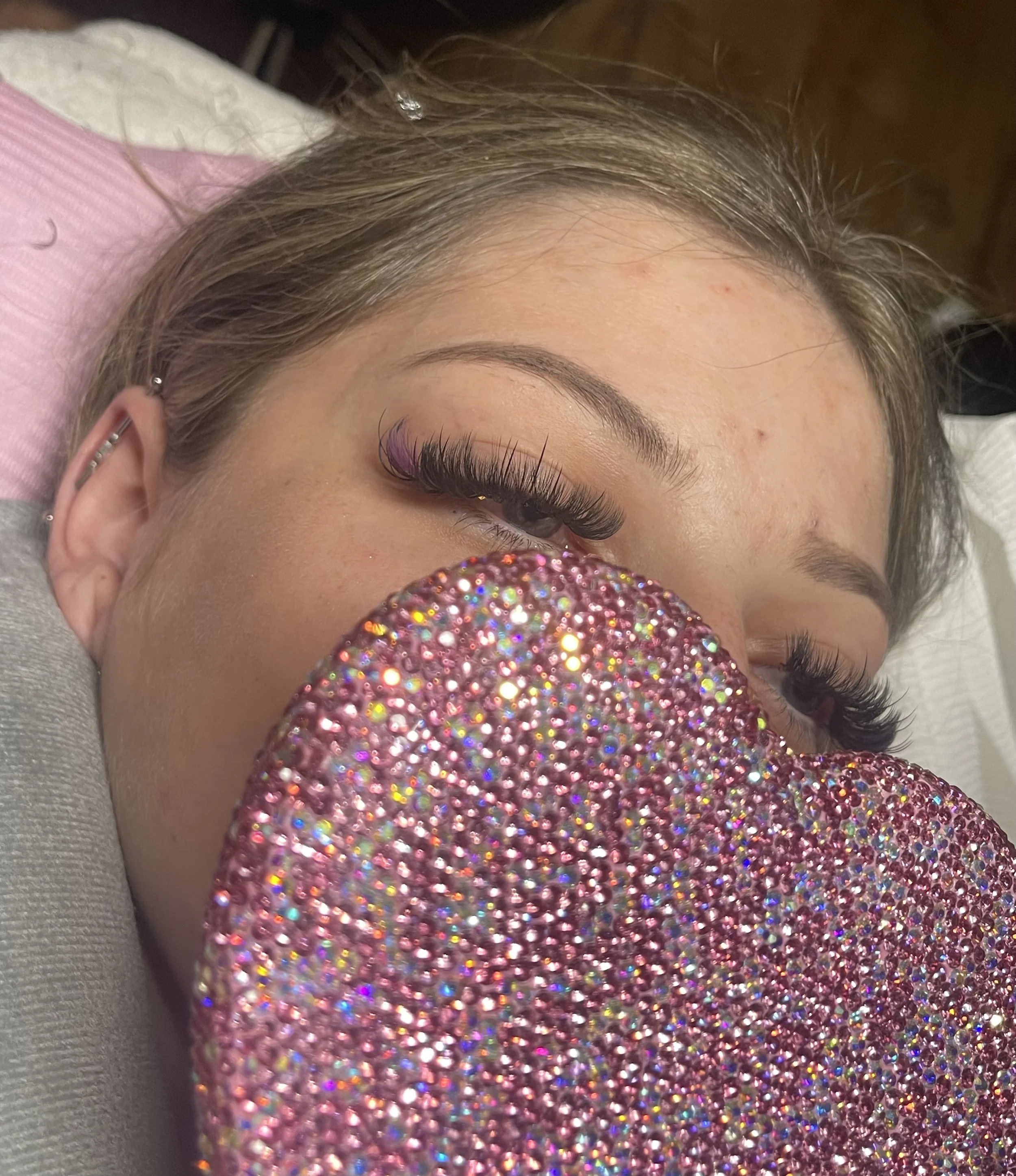 A woman with eyelash extensions lying down, holding a sparkly pink clutch in front of her face.