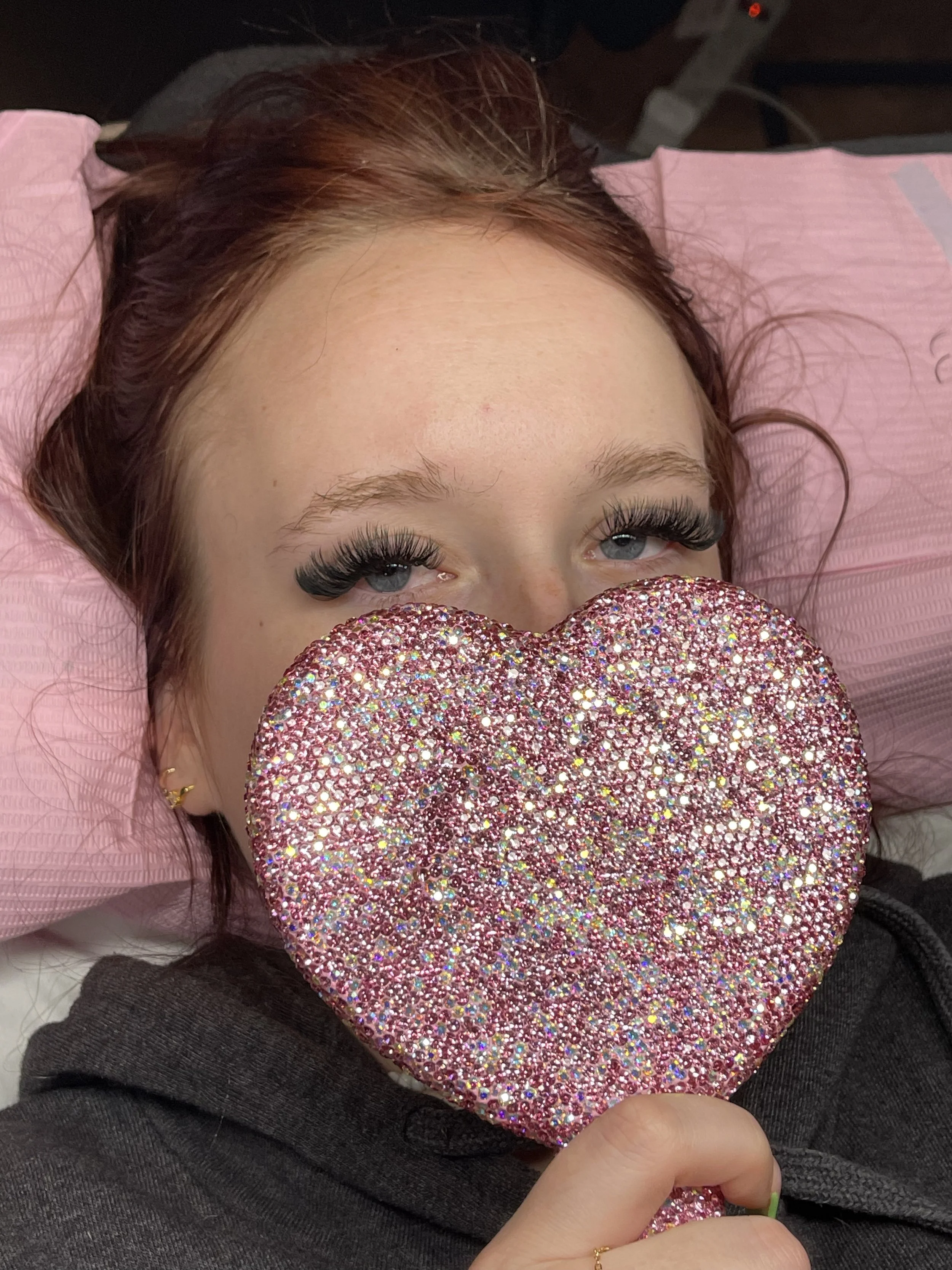 A young woman with red hair and blue eyes, wearing false eyelashes, lies on a pink pillow and holds a large, sparkly pink heart-shaped object in front of her face. She has a small gold earring and is wearing a dark hoodie.