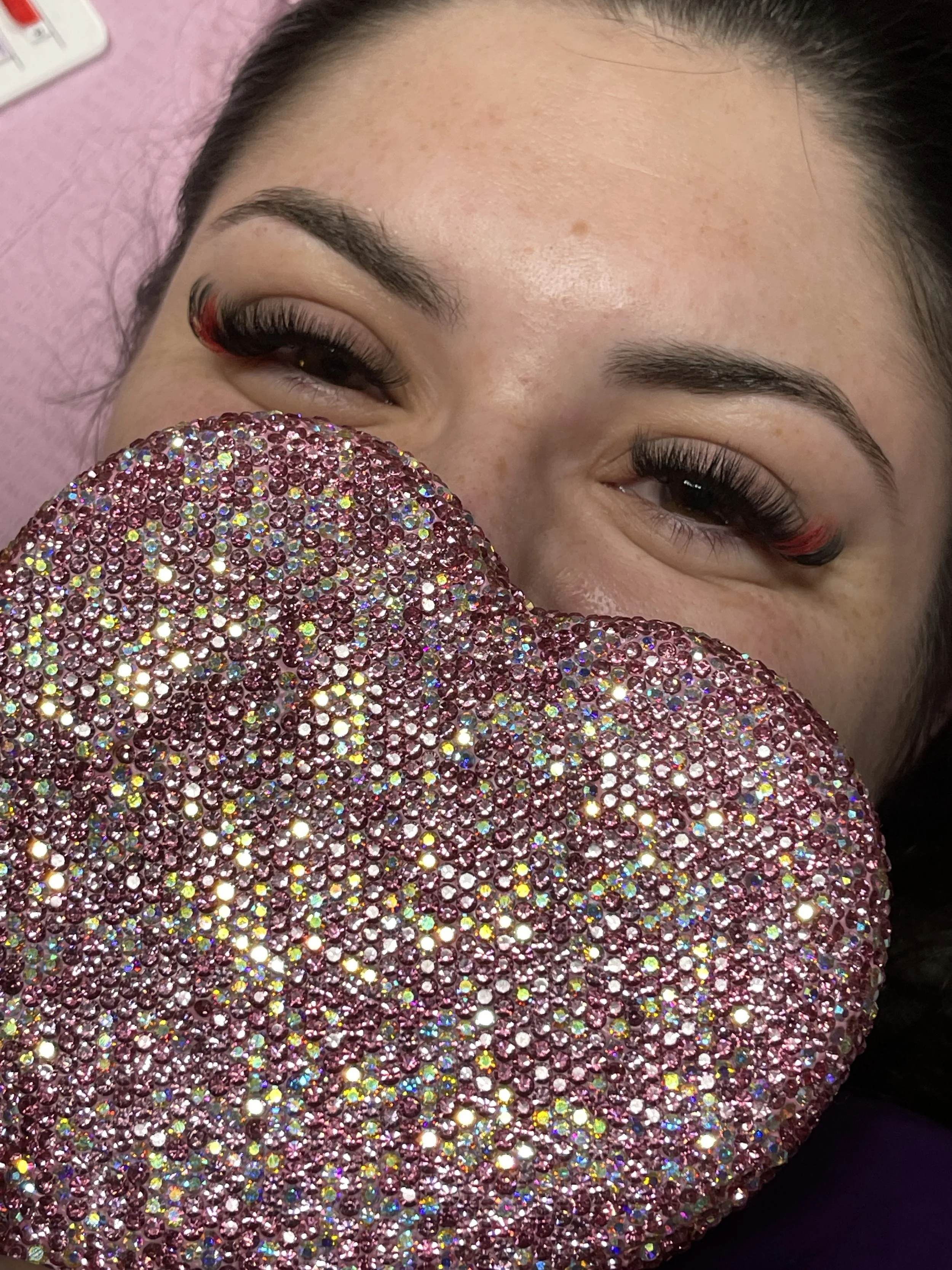 A woman with makeup and long eyelashes holding a large, glittery, pink rhinestone-covered heart in front of her face.