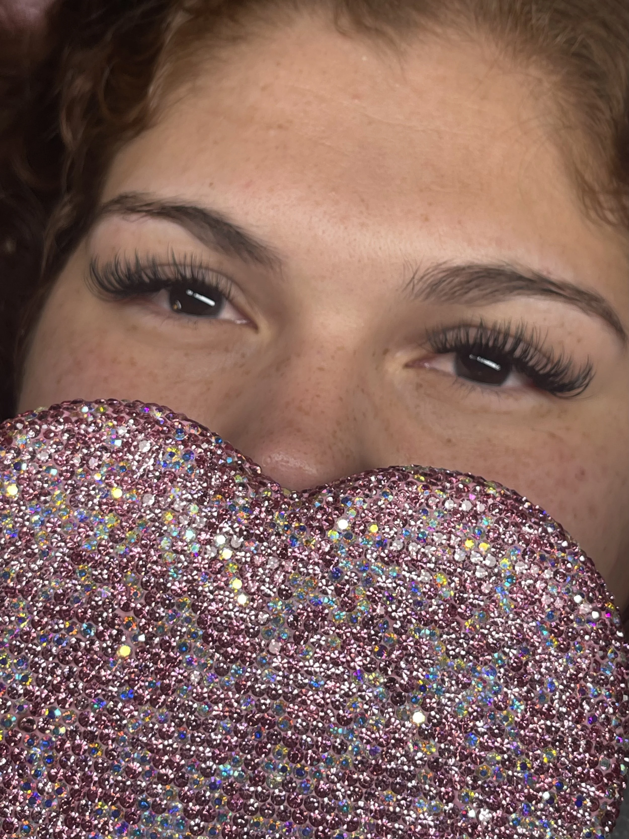 Close-up of a woman's face with freckles, looking over a large, heart-shaped object covered in pink and iridescent rhinestones.