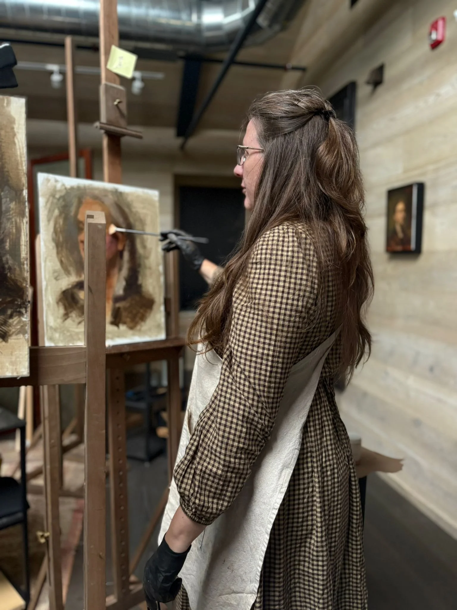 Tanya Stovold with long brown hair, wearing glasses, a checkered dress, and gloves, is painting in a wooden art studio. She is standing in front of an art piece on an easel, focusing on her work.
