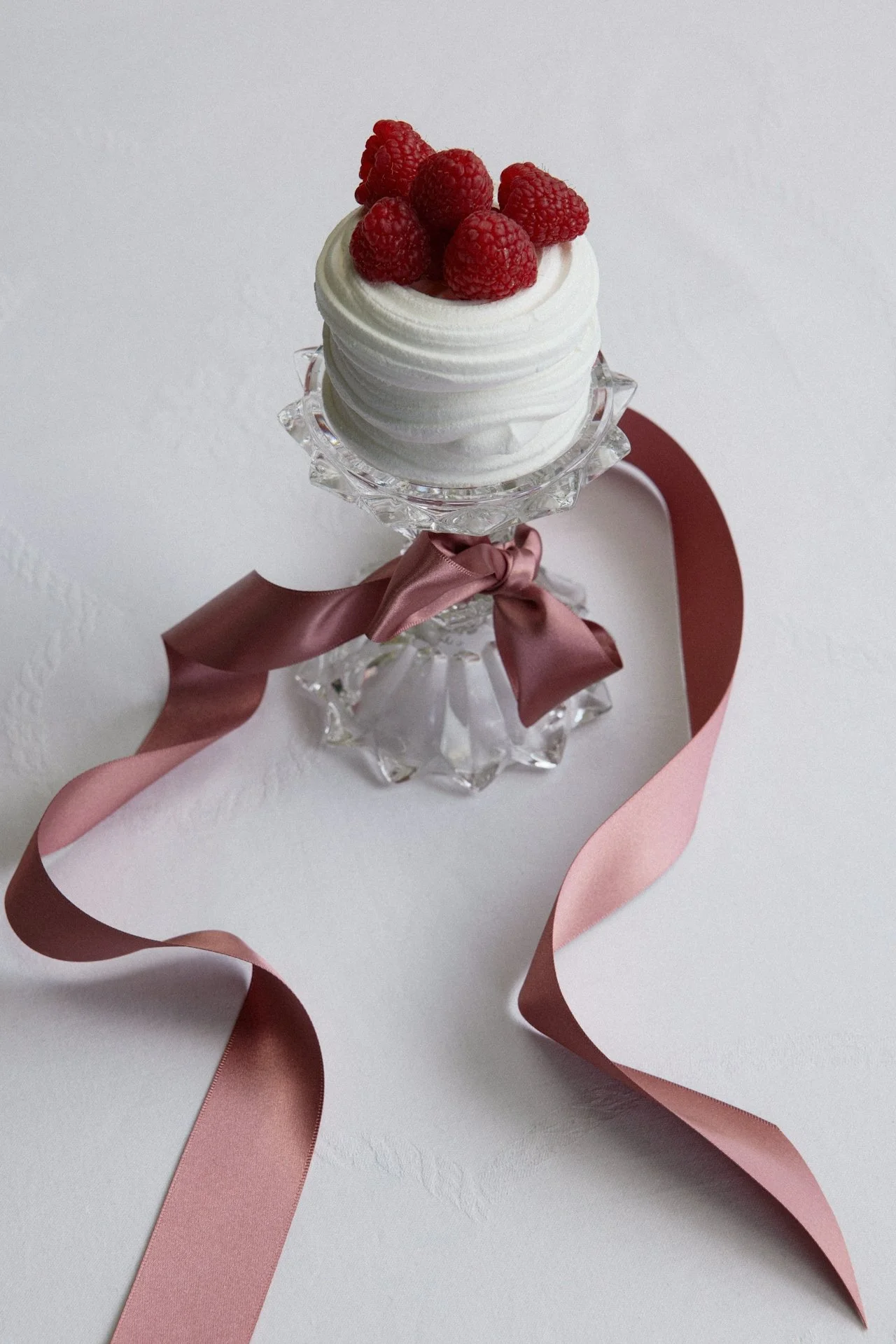 A glass cake stand with a pink ribbon bow holding a small chocolate cake topped with white frosting and fresh raspberries, placed on a white surface with a pink satin ribbon.