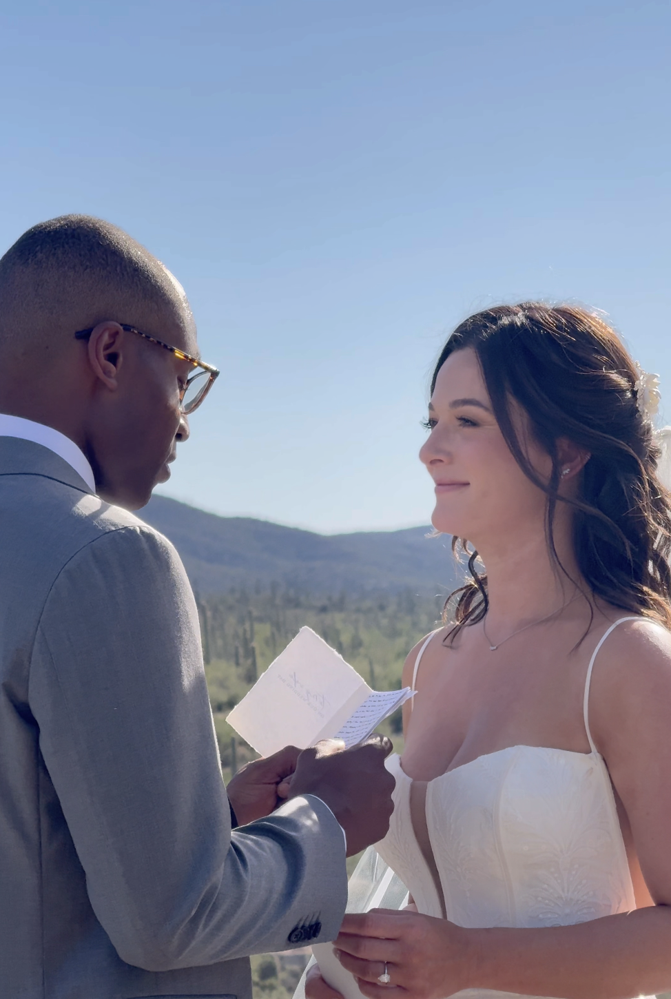 A couple gets married outdoors under a clear blue sky, with mountains in the background. The groom reads vows from a note, and the bride looks happy and emotional.