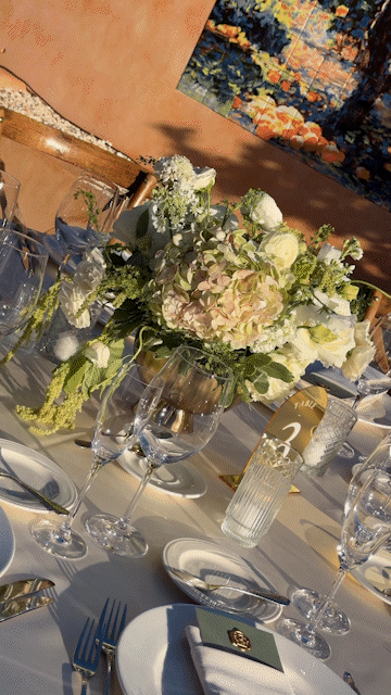 Elegant table setting with a floral centerpiece featuring white and light pink flowers, surrounded by glasses, plates, and silverware, at a formal event or celebration.