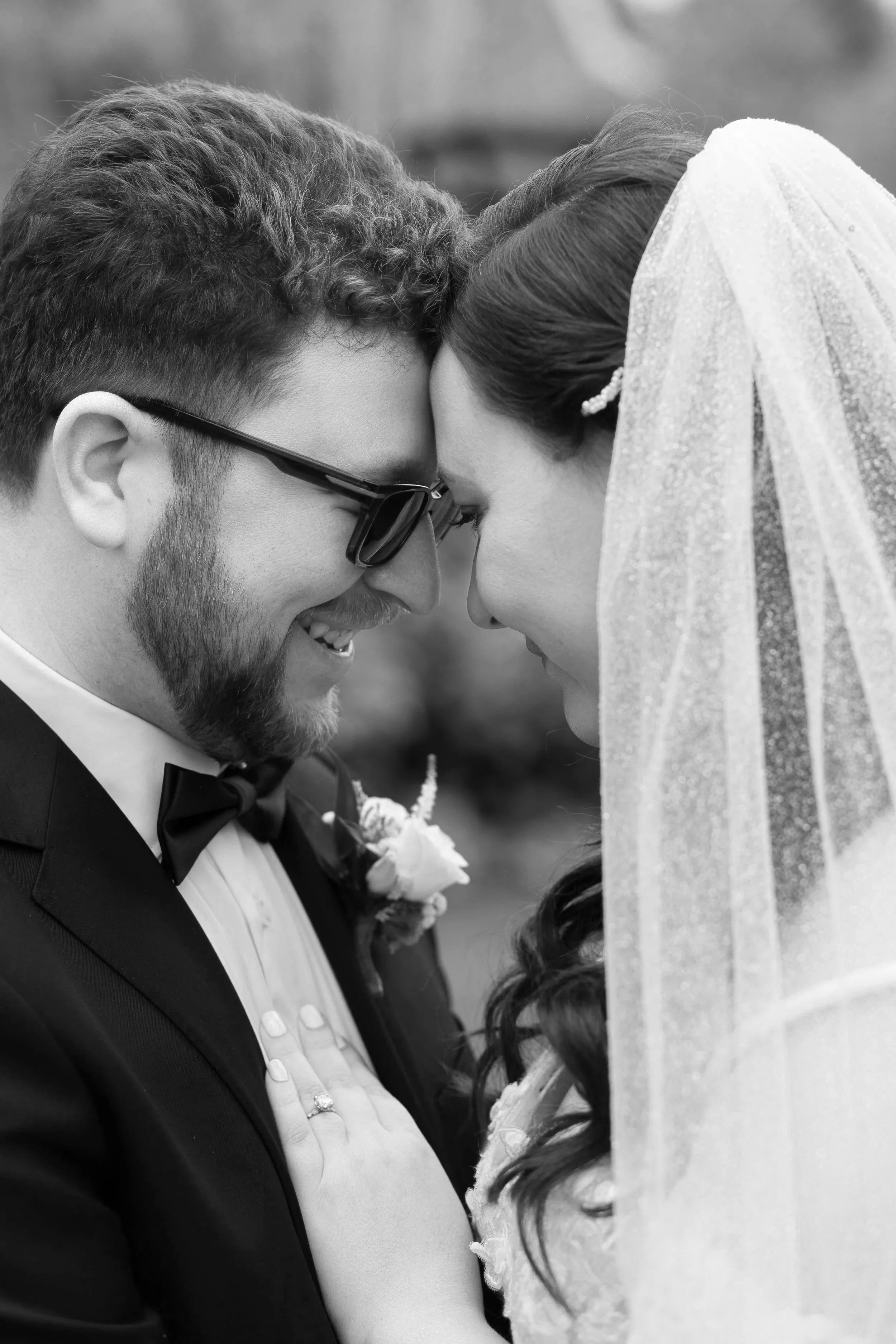 A bride and groom with foreheads touching and eyes closed during a wedding ceremony, with the bride wearing a veil and the groom wearing glasses and a tuxedo.