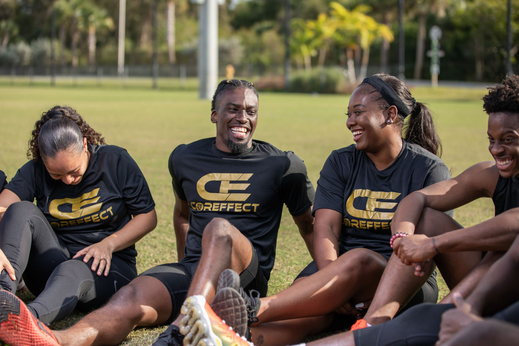 Group of smiling athletes sitting on grass during training, wearing matching black and gold team shirts, outdoors in a park with trees in the background.