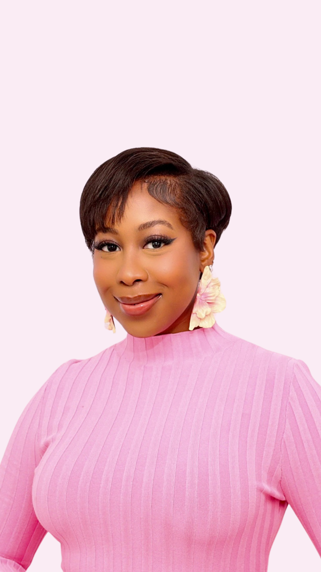 A woman with short dark hair styled in a bob, wearing light makeup, pink lipstick, and large flower-shaped earrings, is smiling and wearing a pink ribbed long-sleeve top.