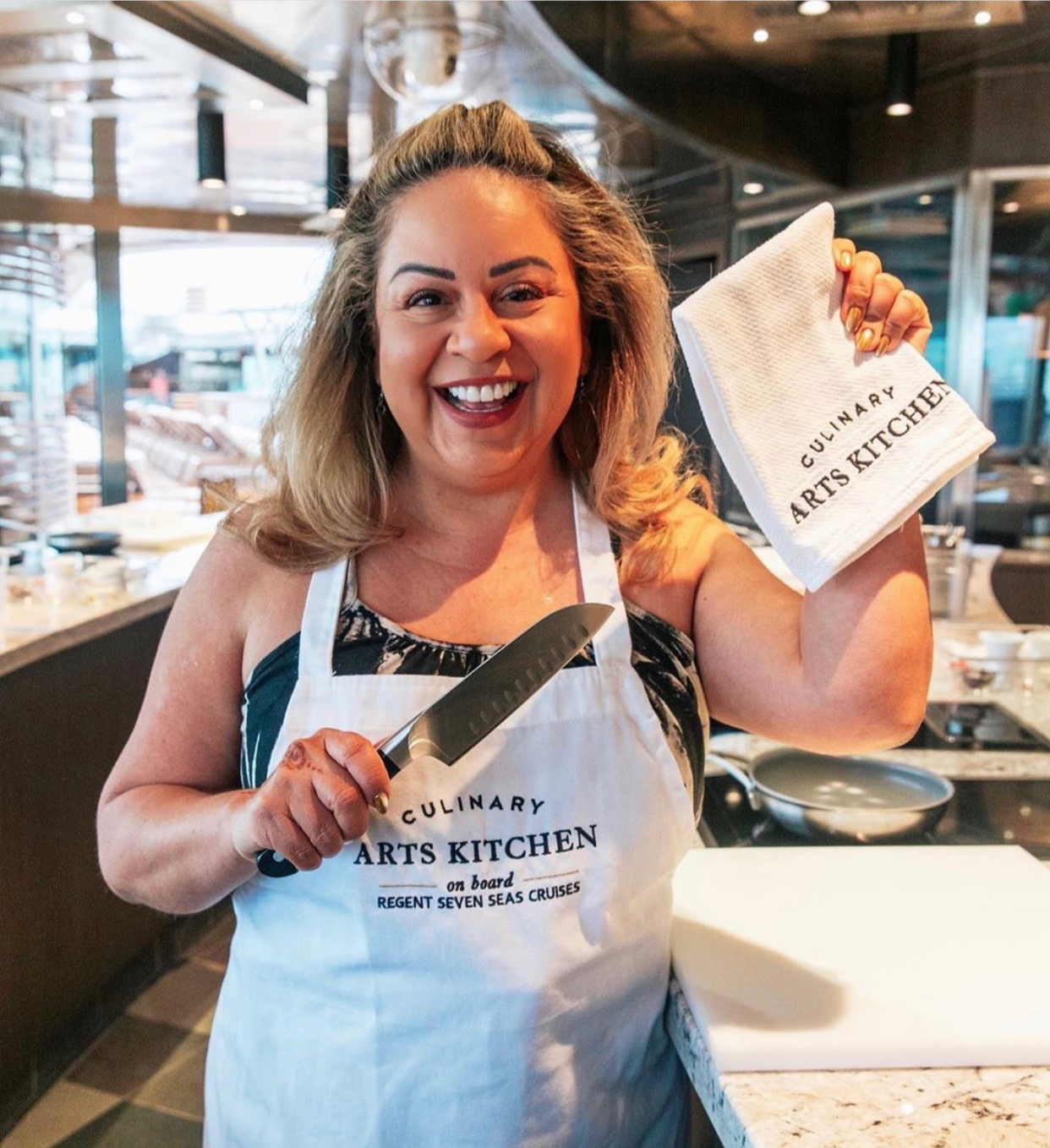 A woman smiling in a kitchen, holding a knife in one hand and a dish towel in the other, wearing an apron that says 'Culinary Arts Kitchen' and 'on board Regent Seven Seas Cruises'.