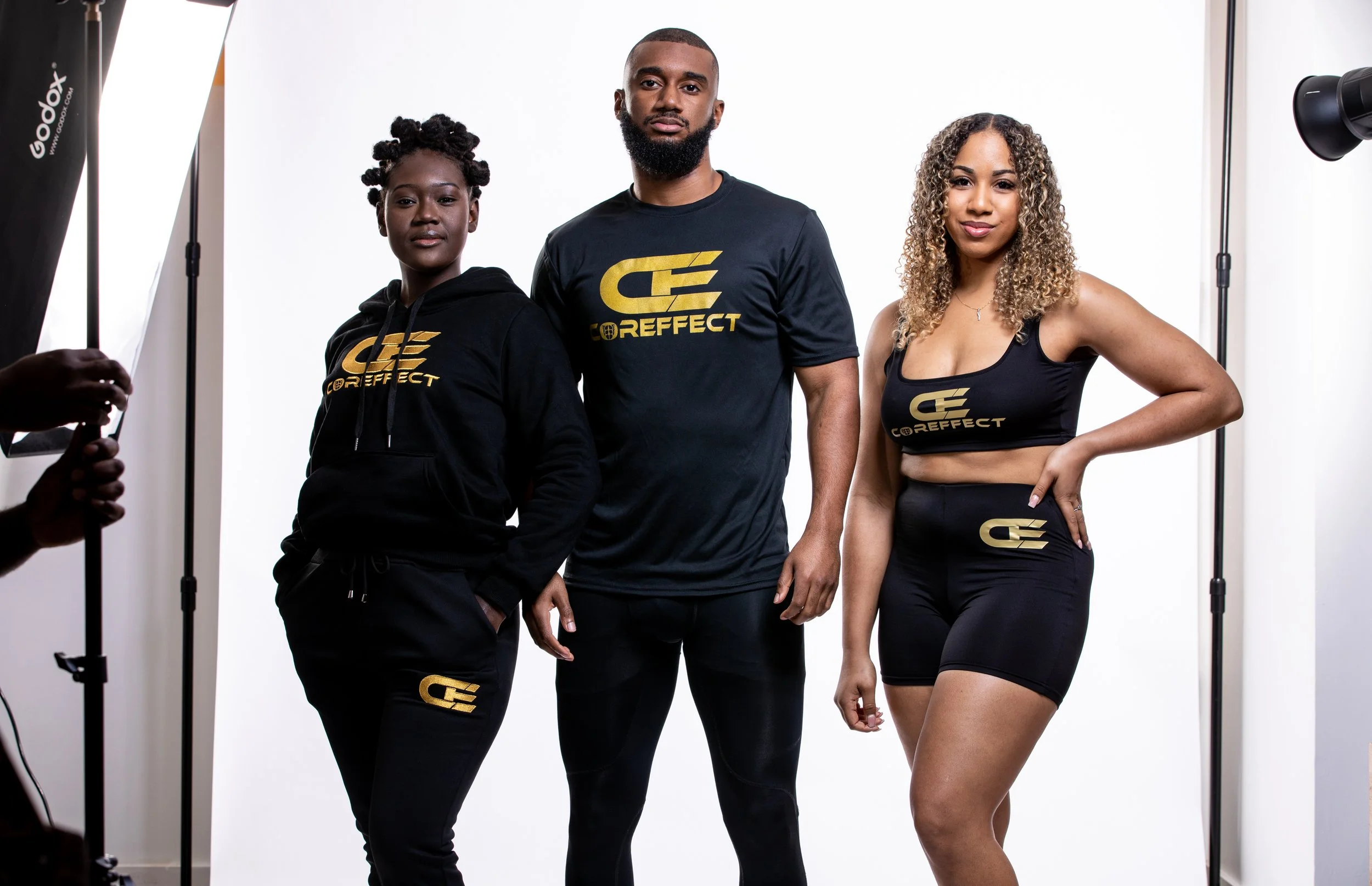 Three individuals posing in front of a white backdrop, wearing matching black workout apparel with gold logo and text, for a photo shoot.