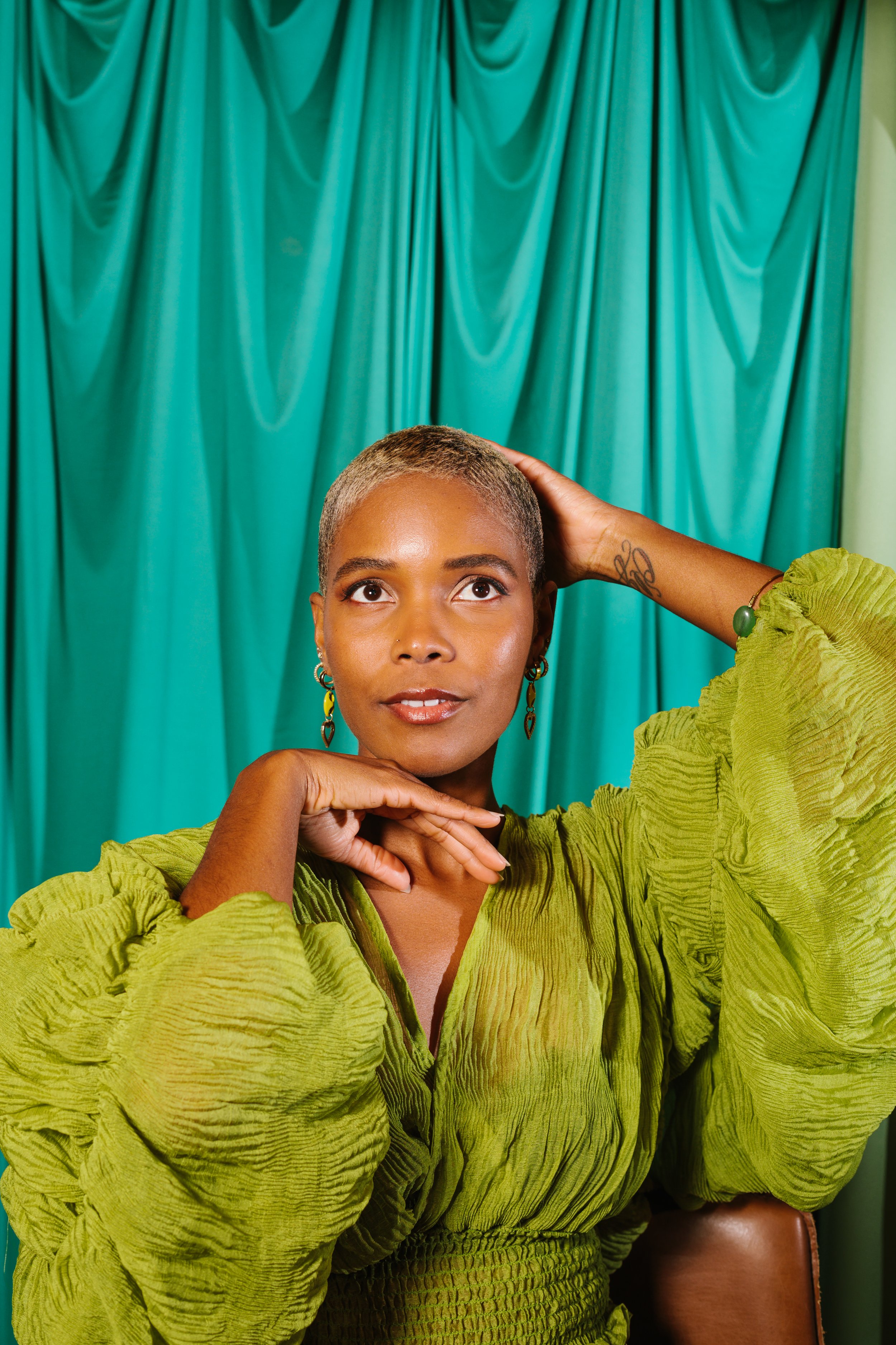 A woman with short, gray hair, wearing a lime green textured blouse, sitting in front of teal curtains, with one hand resting on her neck and the other behind her head, looking upward.