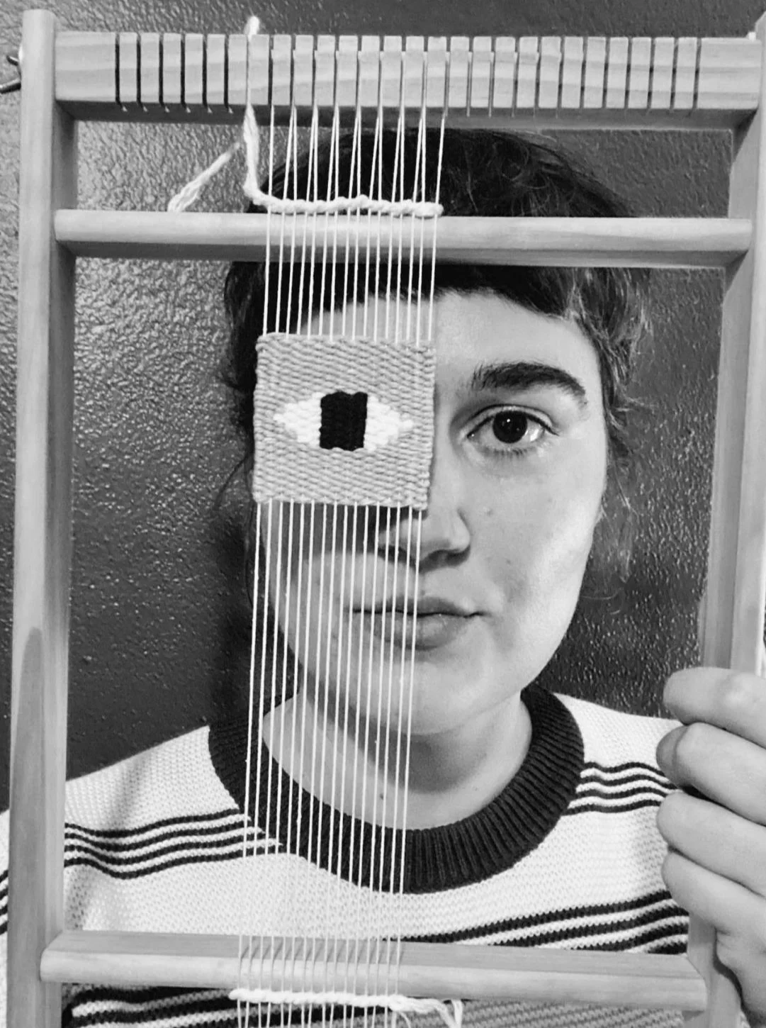 Person holding a wooden weaving frame with a small woven piece depicting an eye, partially covering their face.