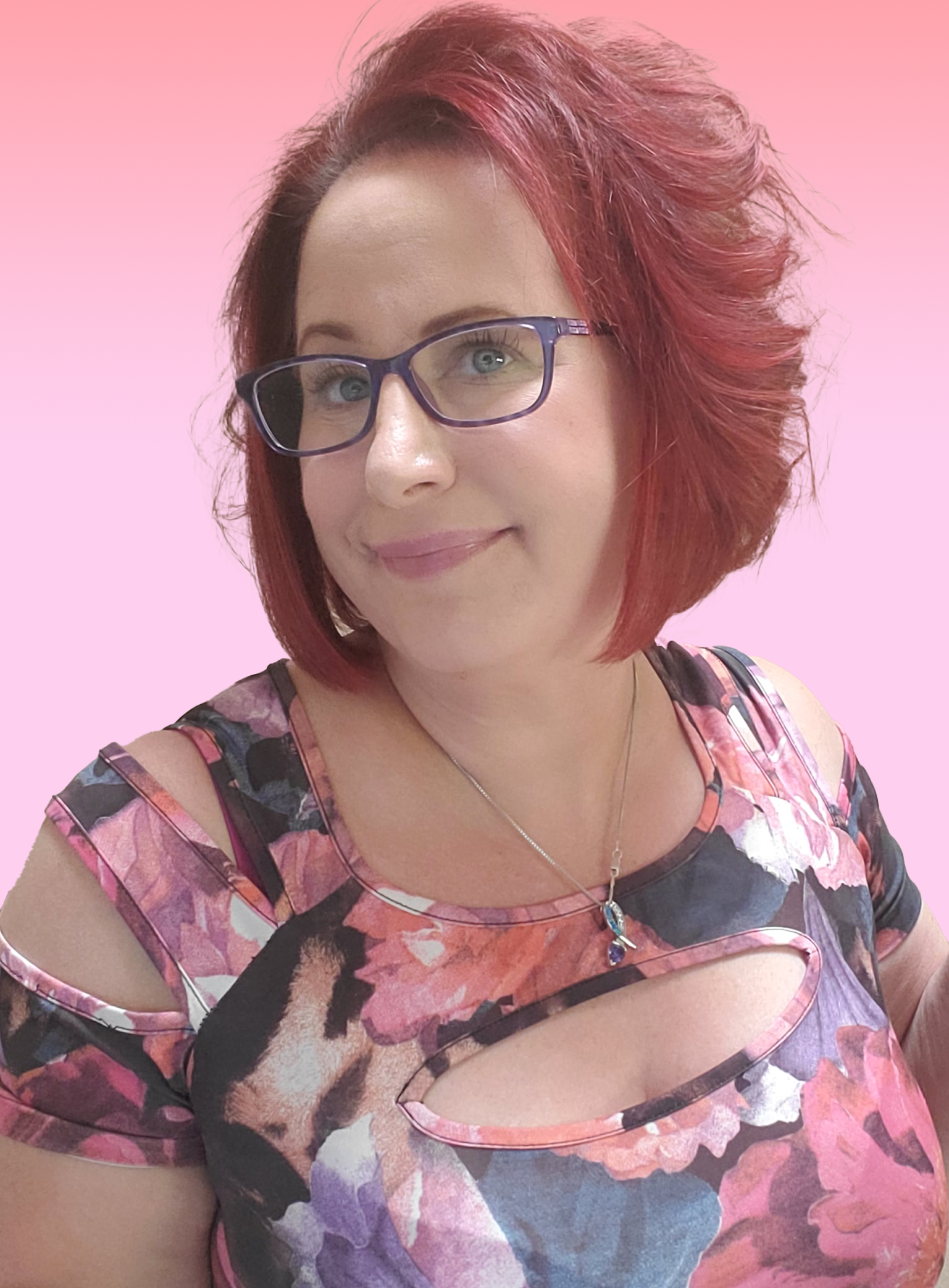 A woman with short, wavy red hair and glasses, smiling, wearing a floral dress with a necklace and pendant, against a pink background.