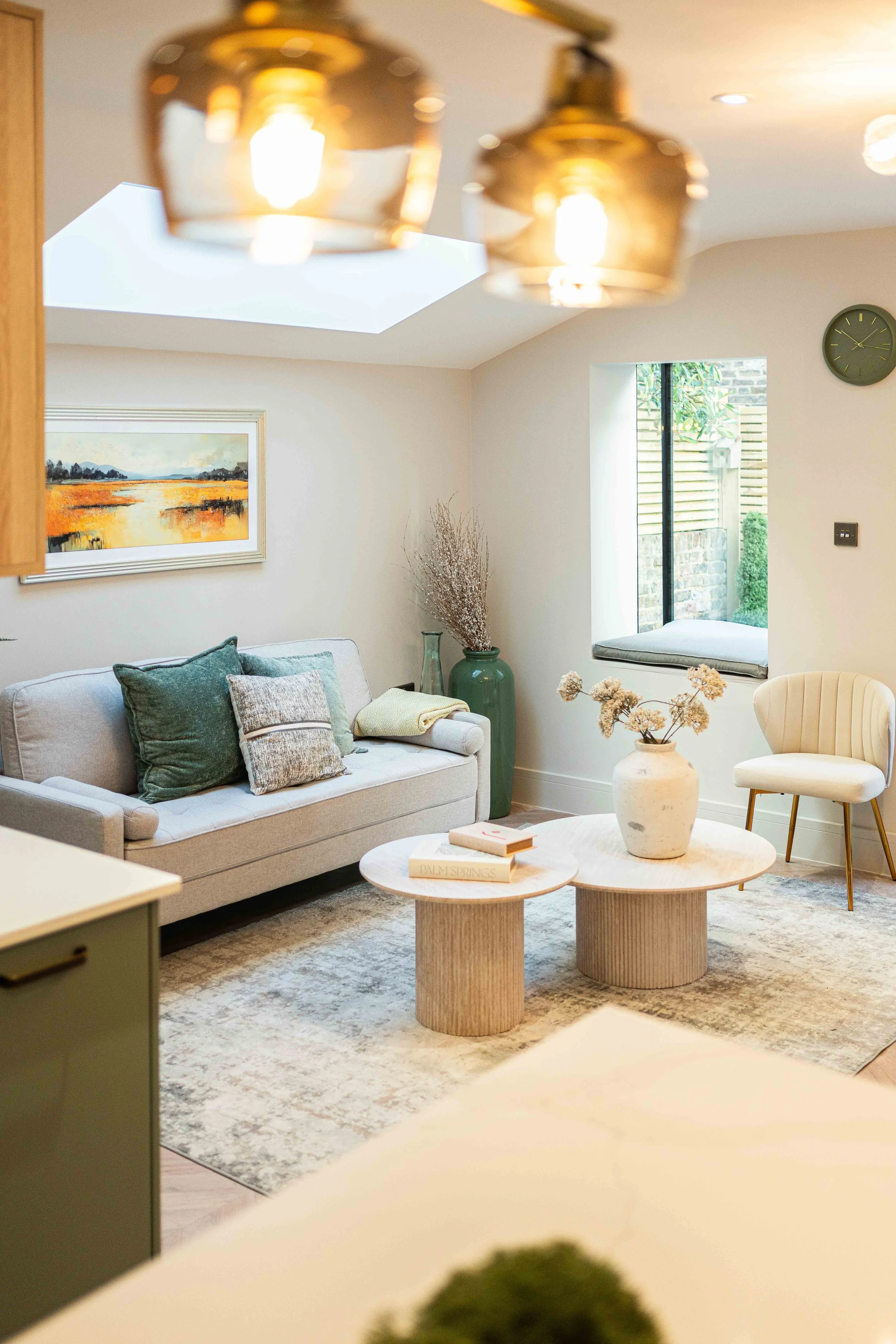 Bright living room with a beige sofa, green and gray pillows, a white armchair with gold legs, two round wooden coffee tables with decorative vases and books, a large window with a cushioned seat, a green vase with dried branches, a wall clock, and a