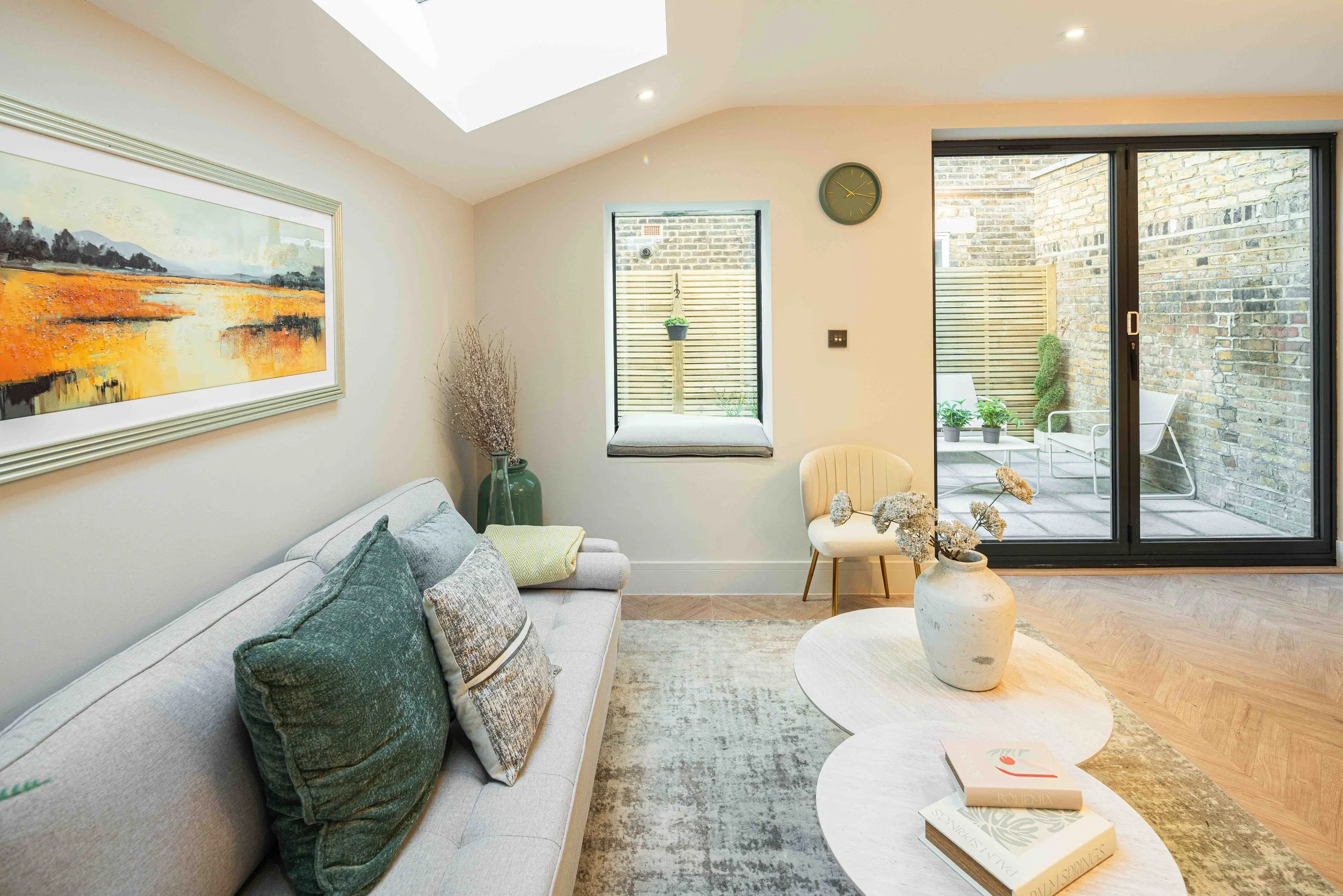 Living room with a light gray sofa, decorative pillows, a large framed landscape painting on the wall, a beige armchair, a white vase with dried flowers, a small coffee table with books, a window seat with a cushion, a large sliding glass door leadin