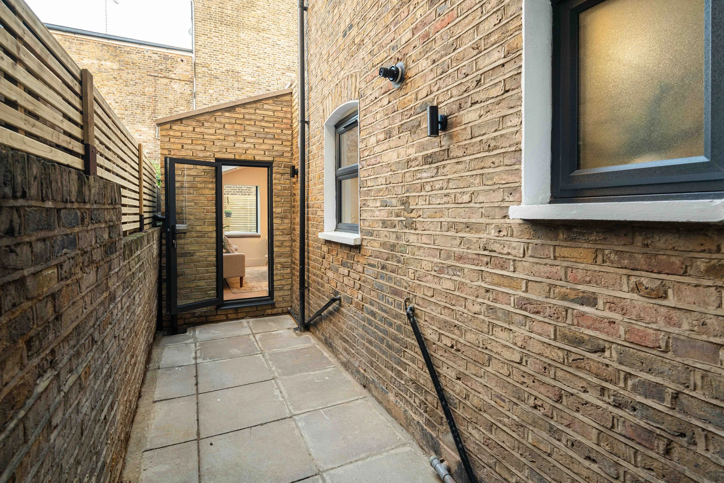 Small outdoor patio area with concrete tiles, bordered by brick walls, leading to a glass door and a window on a brick building