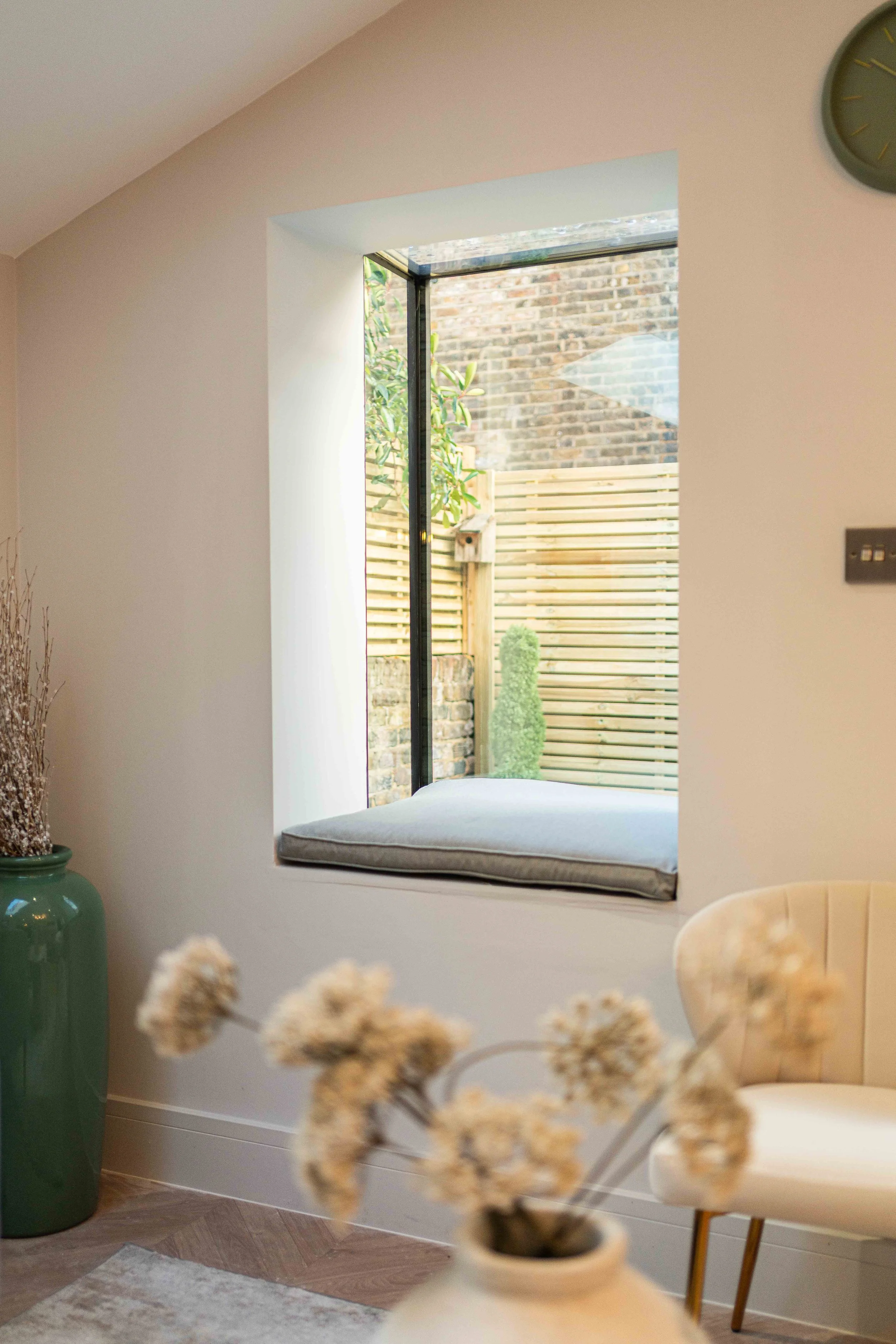 Interior of a room with a large window seat, showing a view of a small garden with a wooden fence and a green shrub outside.