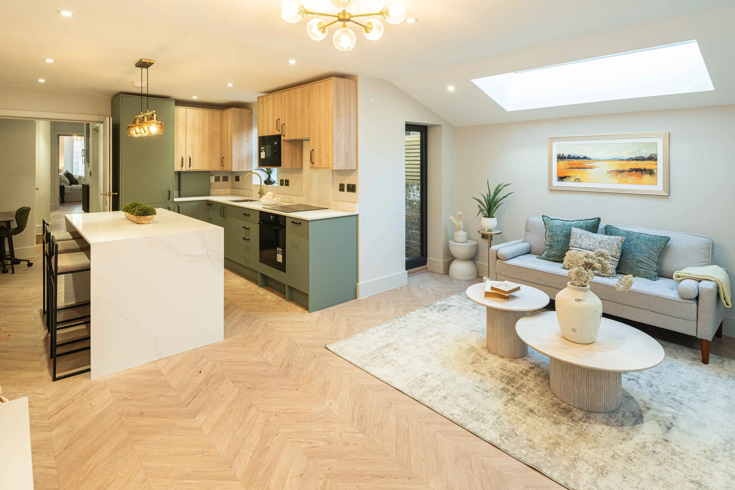 Open-concept living room and kitchen with modern decor, light wood flooring, a gray sofa with pillows, a small round coffee table, a kitchen island, pastel green and wood cabinets, hanging light fixtures, and a large skylight.