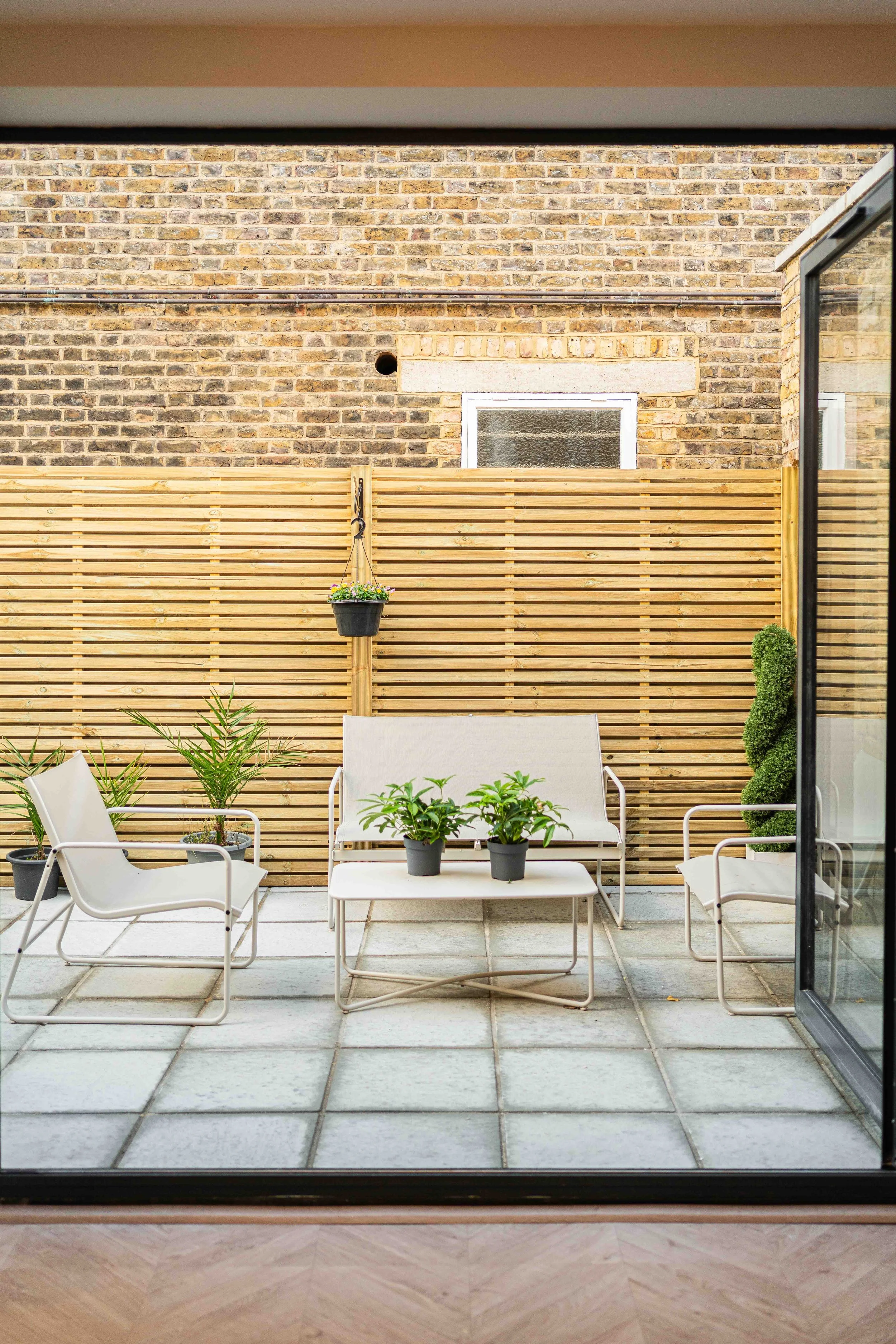 View of a small outdoor patio with white metal chairs, a matching white metal sofa with potted plants on the table, potted plants along the edges, a wooden privacy screen, brick wall, and sliding glass door.