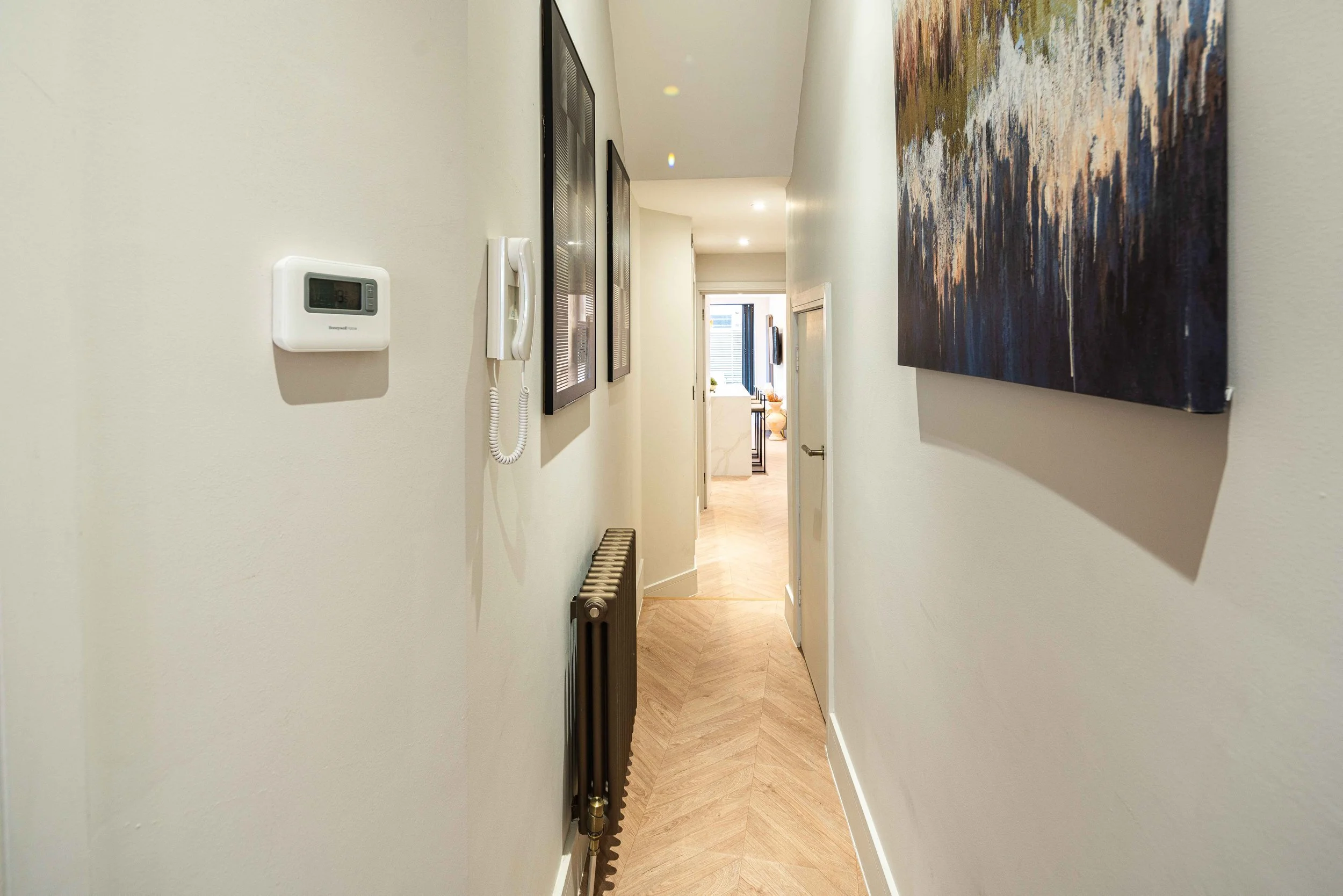Narrow hallway with light-colored walls, hardwood floor, framed artwork, thermostat, wall-mounted phone, and radiator, leading to a brighter room at the end.