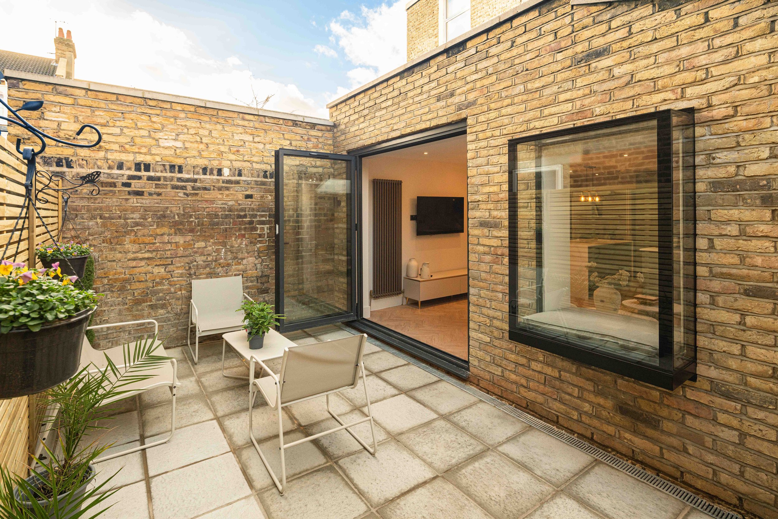 Small rooftop patio with beige stone tiles, outdoor furniture including three white chairs and a small table with a potted plant, brick wall enclosure with hanging flower pots, open glass door leading to a living room with wall-mounted TV, large wind