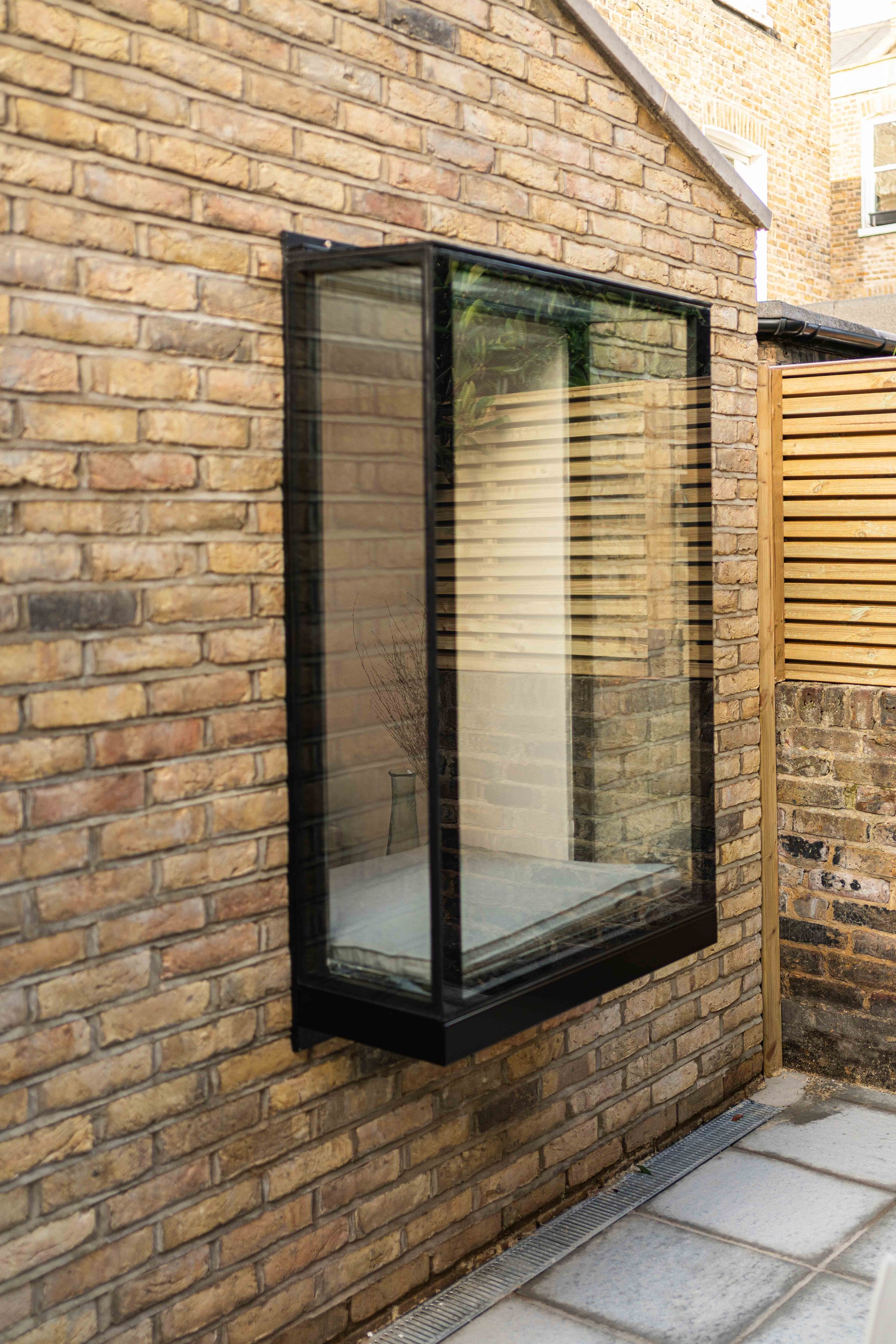 A rectangular glass cabinet with a black frame mounted on a brick wall outside a building, reflecting nearby greenery and part of the brick wall.
