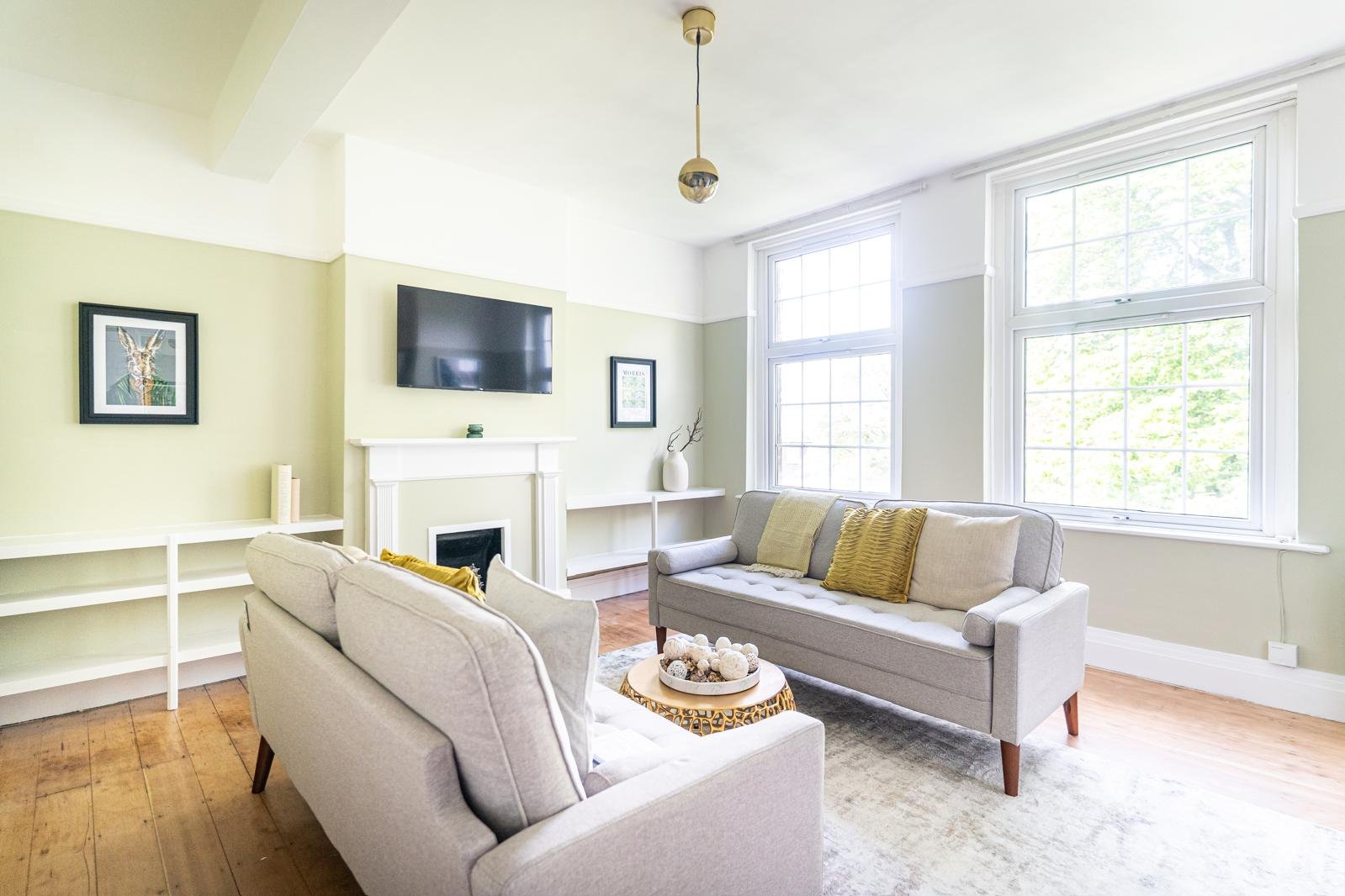 Bright living room with two beige sofas, large windows with white trim, a black flat-screen TV mounted above a white fireplace, decorative pillows on the sofas, a round coffee table with decor, a white built-in shelf, pale green walls, wooden floor, 