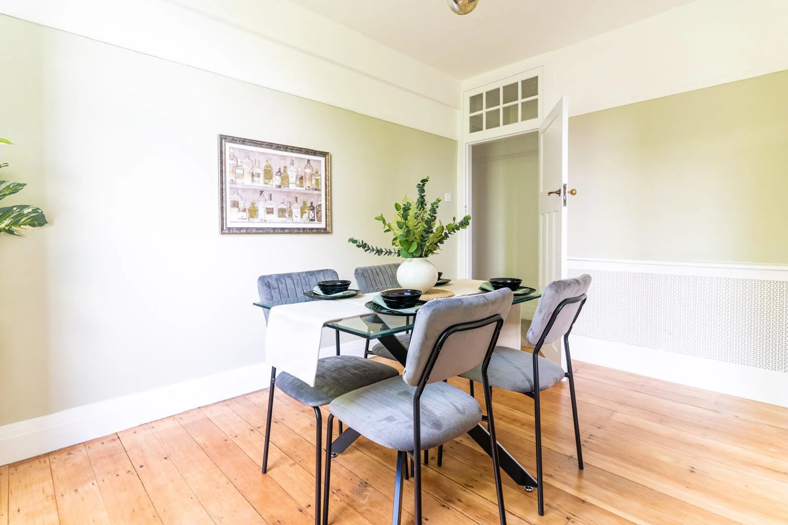 Dining room with a rectangular glass table, six grey upholstered chairs, a white vase with green foliage, a framed picture of bottles on the wall, hardwood floors, and a partially open door.