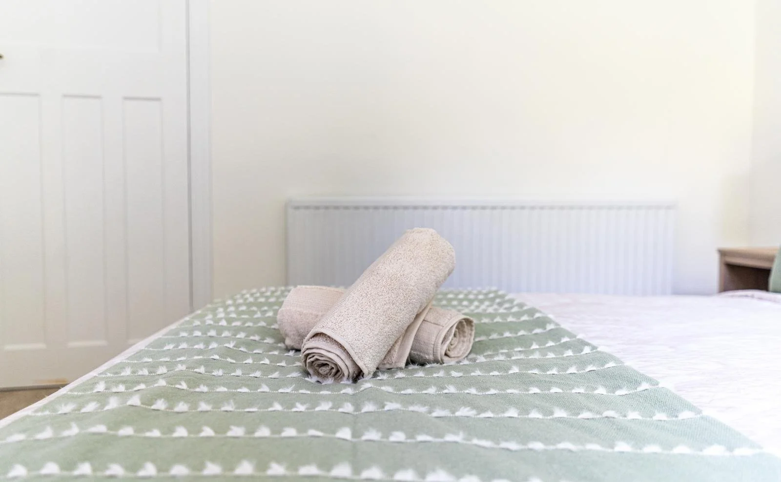 A neatly made bed with rolled towels placed on a green and white patterned bedspread in a dormitory or hotel room with a white door and radiator in the background.