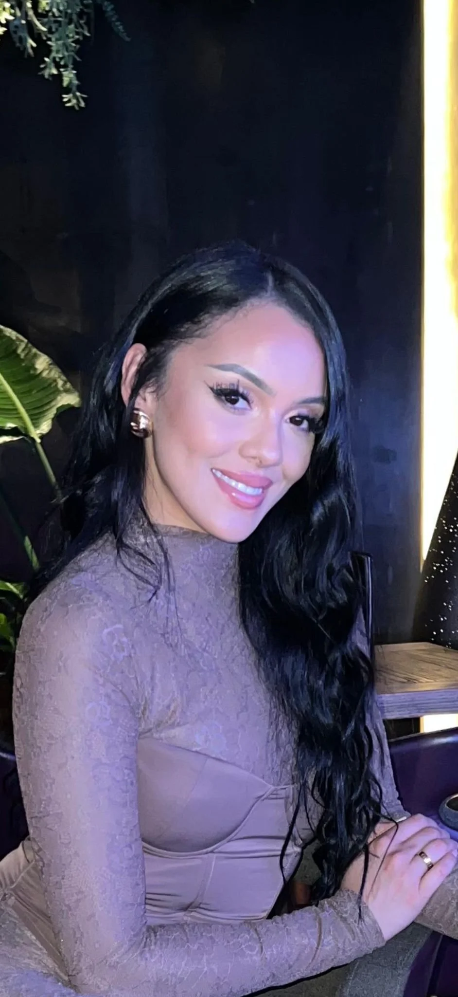 A woman with long black wavy hair, wearing gold earrings and a beige lace long-sleeve dress, smiling at the camera in a dimly lit setting with green plants and a dark background.