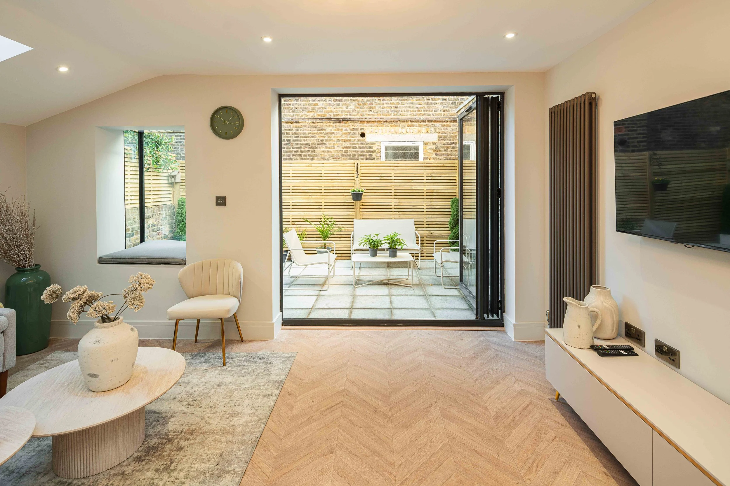 Living room with light wood flooring, a white coffee table with plant arrangements, a beige armchair, a white media console with decorative vases, a wall-mounted TV, a window seat with a cushion, and a sliding glass door leading to a patio with outdo