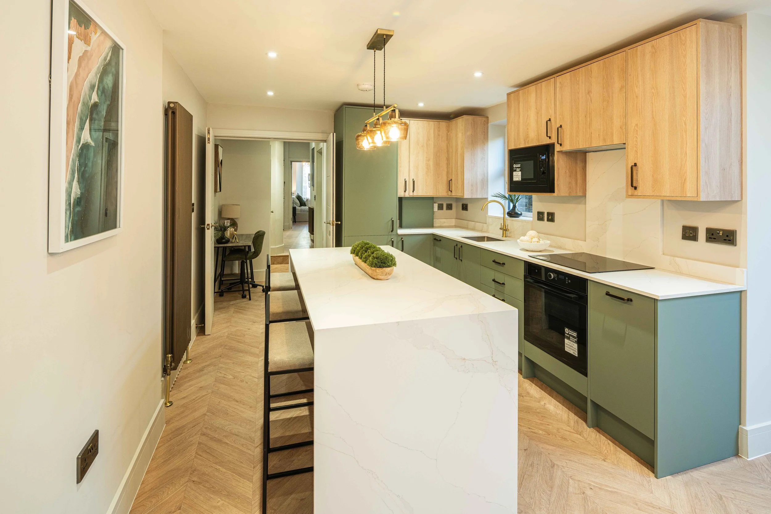 Modern kitchen with green lower cabinets and wooden upper cabinets, white marble countertop island, black built-in appliances, gold faucet, small window with plant, pendant lights, and a hallway leading to a living room.