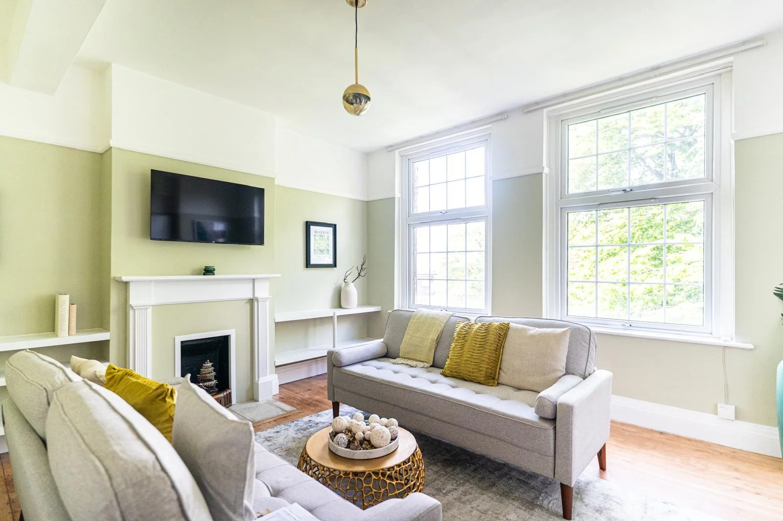 Bright living room with large windows, light green walls, a wall-mounted TV above a white fireplace, two light-colored sofas with yellow and beige pillows, a round wooden coffee table with decorative balls, and minimal decor including a framed pictur