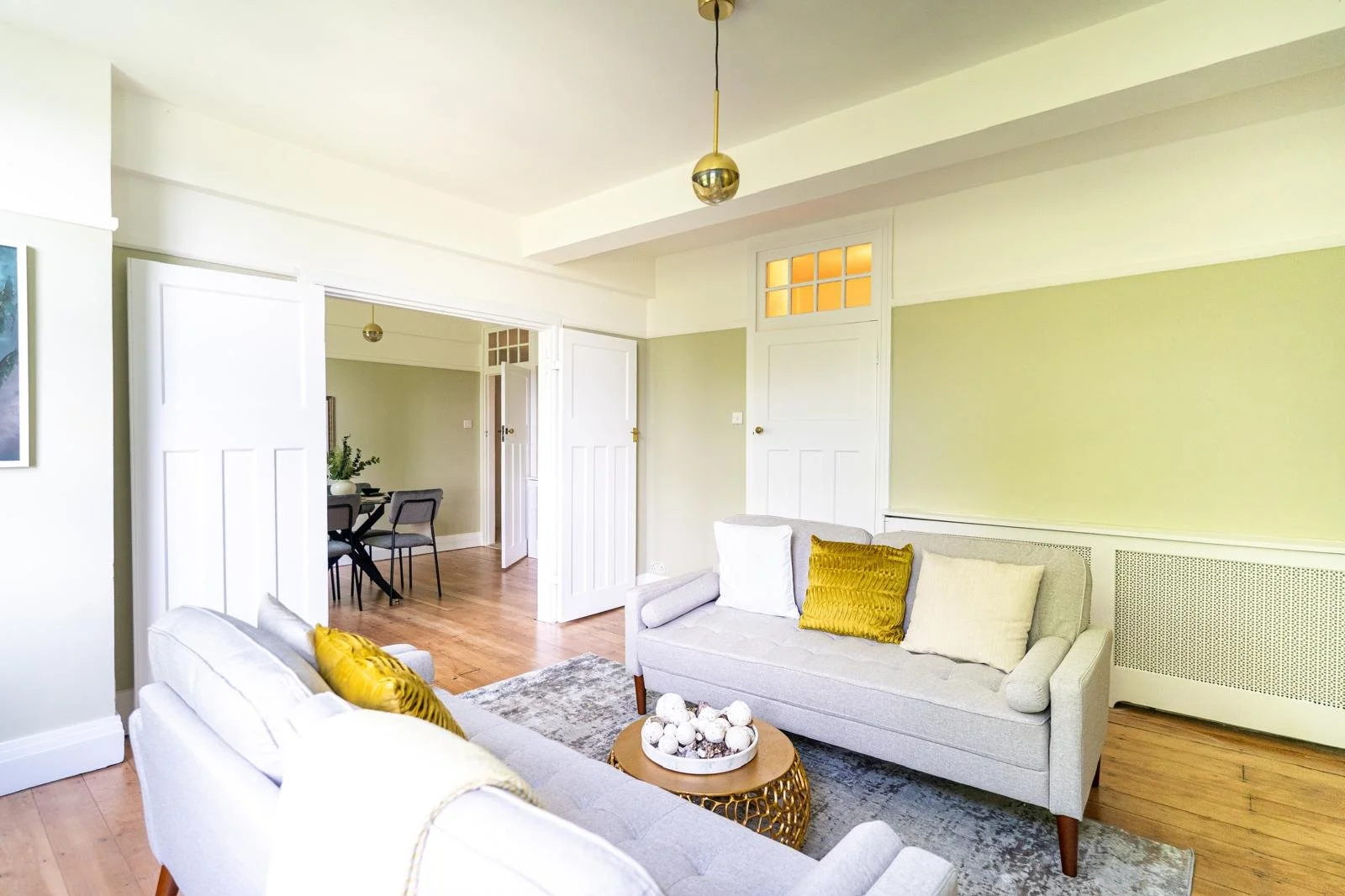 Bright living room with light-colored sofas, yellow and cream pillows, a wooden coffee table with decorative balls, hardwood floors, and an open doorway leading to a dining area.