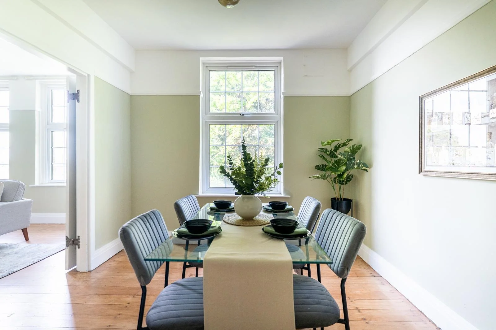 Dining room with a glass table, six gray upholstered chairs, a vase with greenery, and a window.