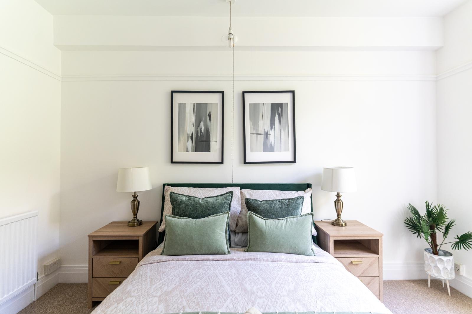 A neatly made bed with green and white pillows, two wooden nightstands with white lamps, abstract framed art on a white wall, and a potted plant in a white pot on the right side.